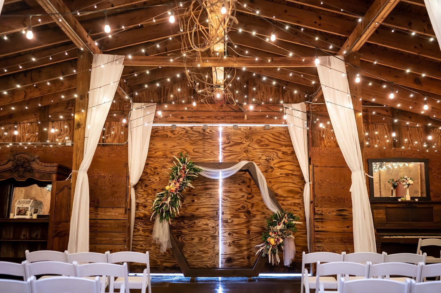 Rustic barn wedding venue interior with string lights, white drapes, and greenery decorations against wooden walls.