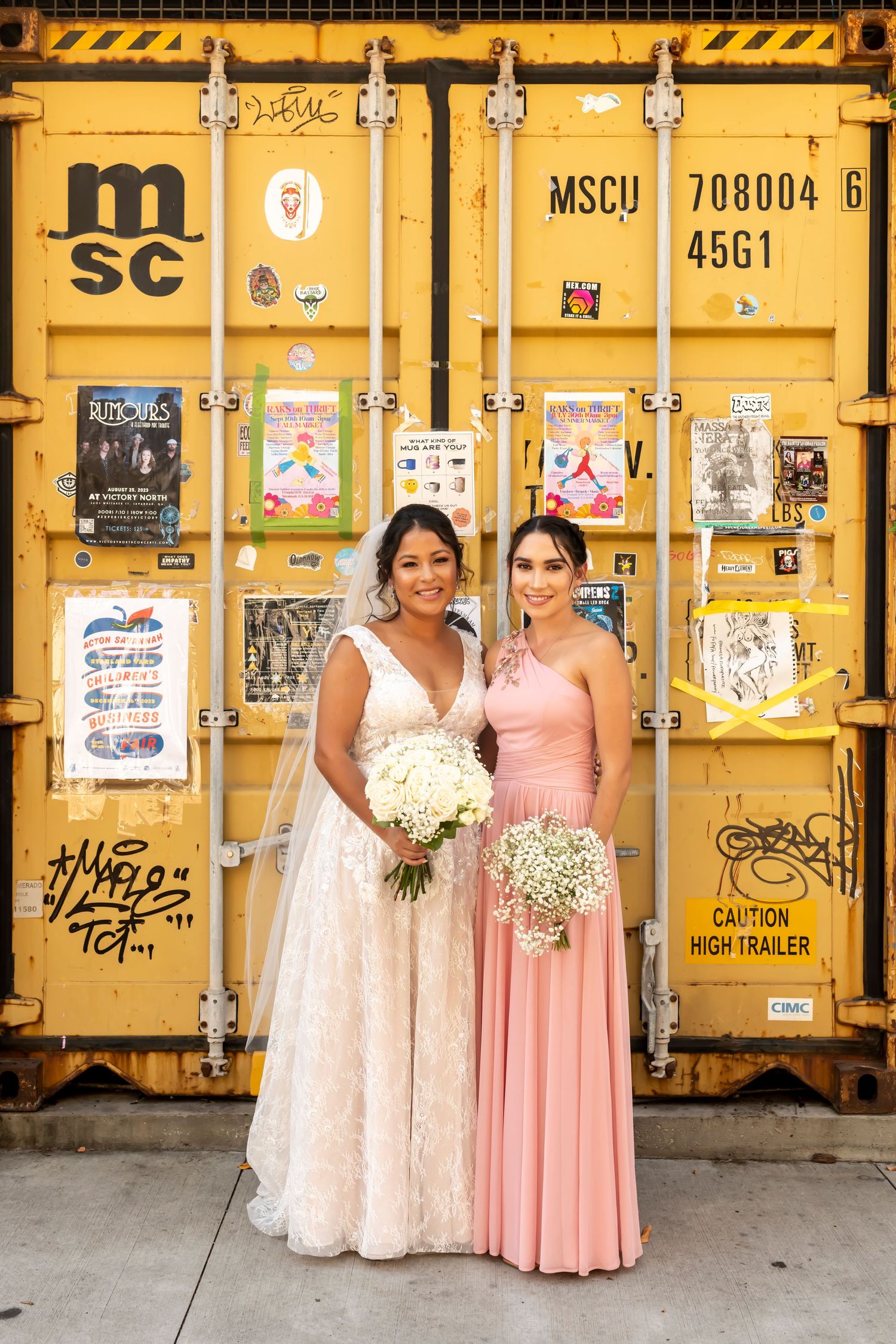 Two attendants in pink and cream formal dresses pose next to yellow shipping containers with graffiti and signage.