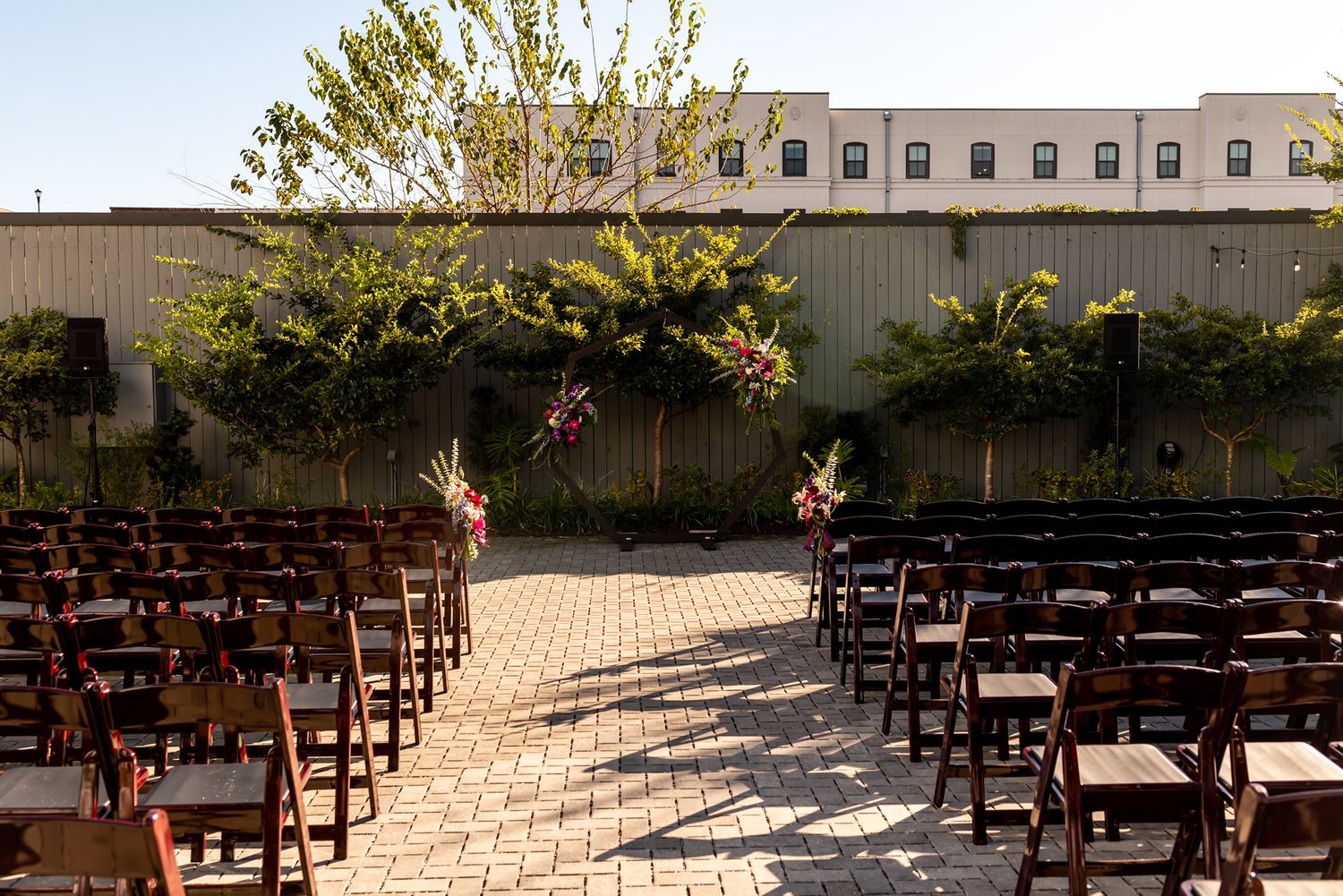 Rows of wooden chairs arranged for an outdoor ceremony on a stone patio with trees and buildings in the background.
