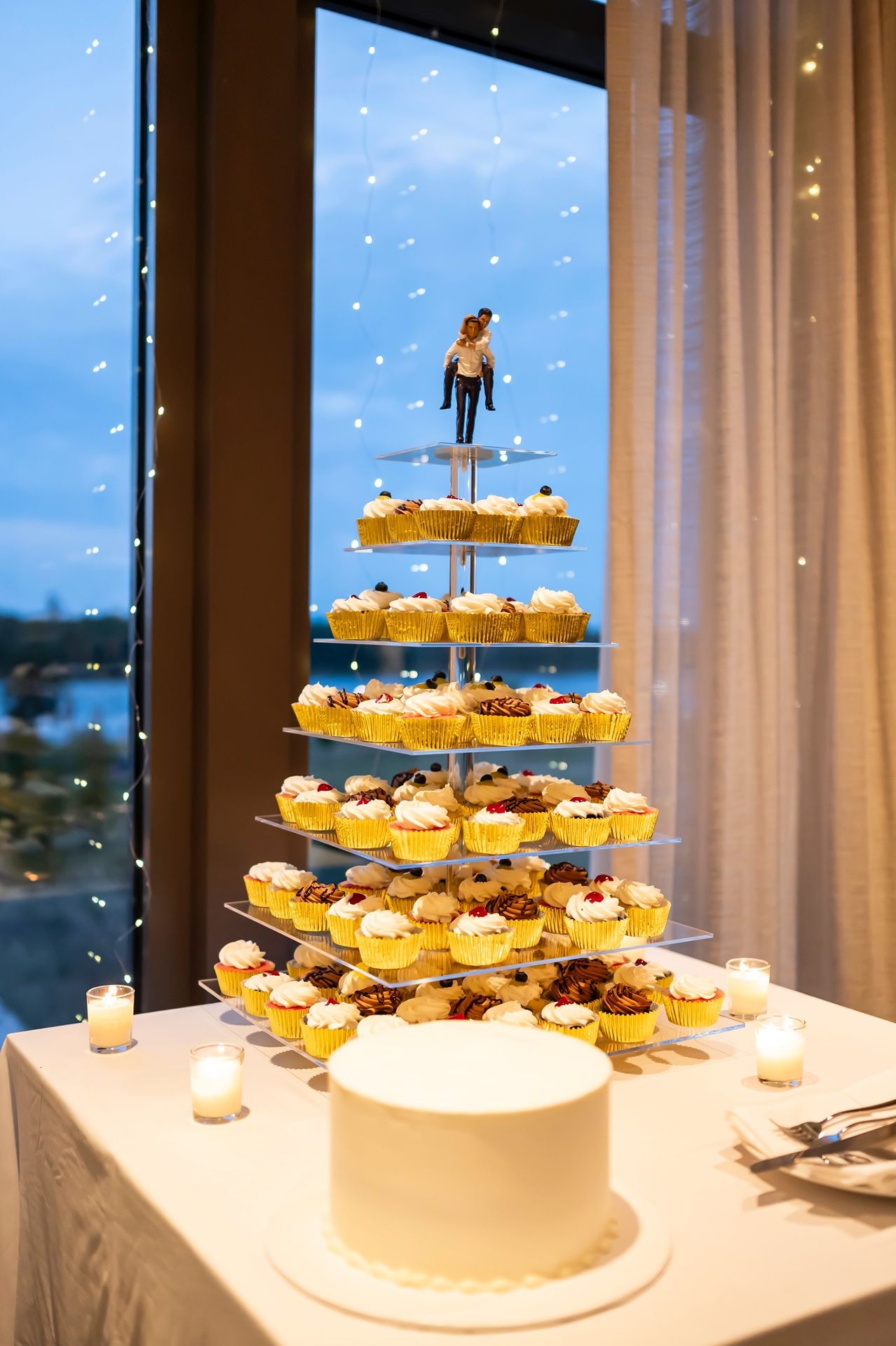 A wedding cake display featuring a tiered stand of cupcakes topped with a cake topper, set against a window at dusk.