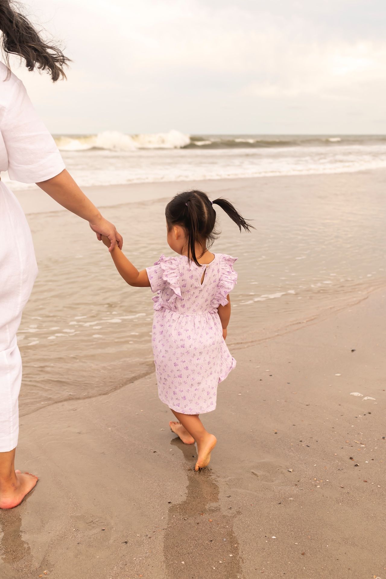 Parent and young child holding hands while walking barefoot along a misty beach shore in matching pink and white outfits.