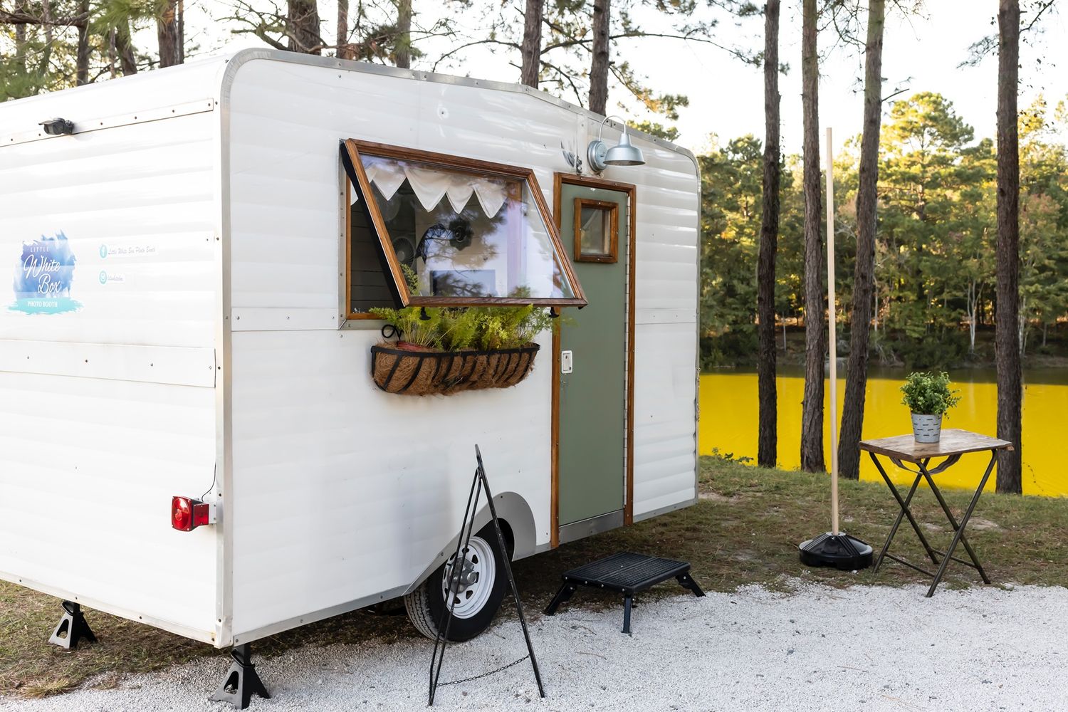 Small white vintage trailer with window flower box and door parked in snowy pine forest at sunset.
