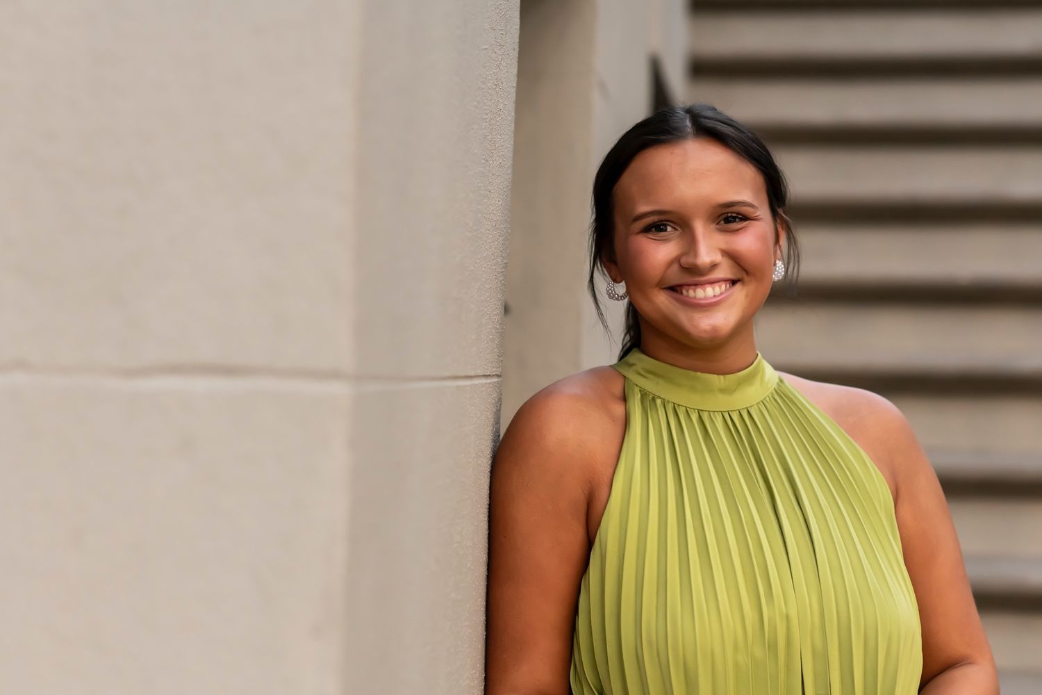 A person in a chartreuse pleated halter top smiles warmly while standing against a light-colored wall.
