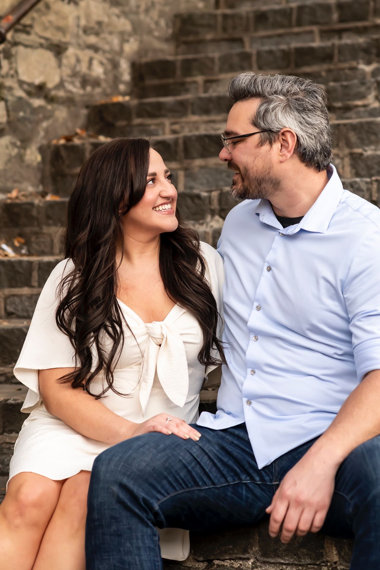 A couple sits together on stone steps sharing a candid laughing moment against a rustic brick wall background.