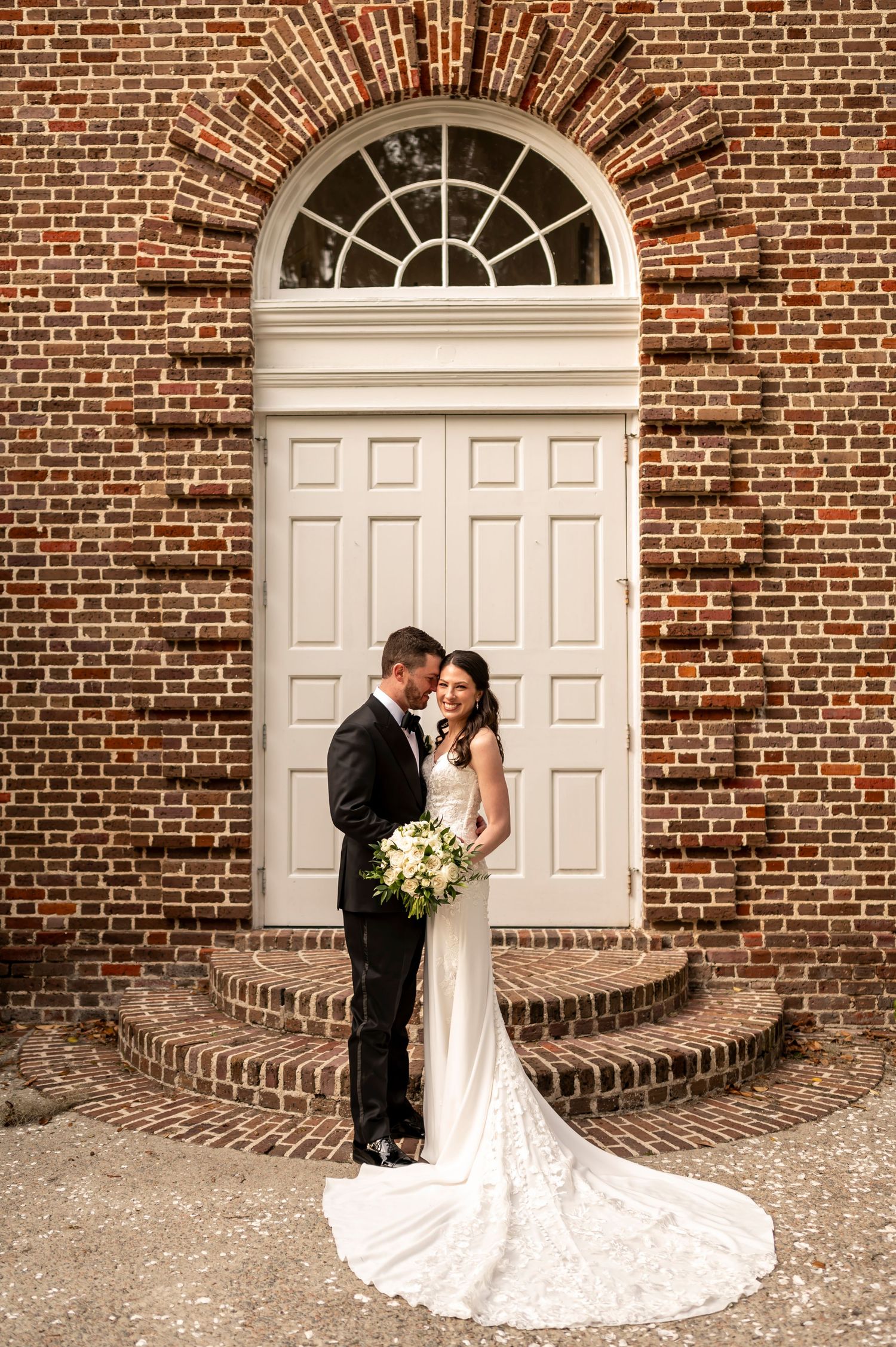 Elegant couple shares intimate moment in front of grand arched brick doorway with curved transom window and sweeping bridal train.
