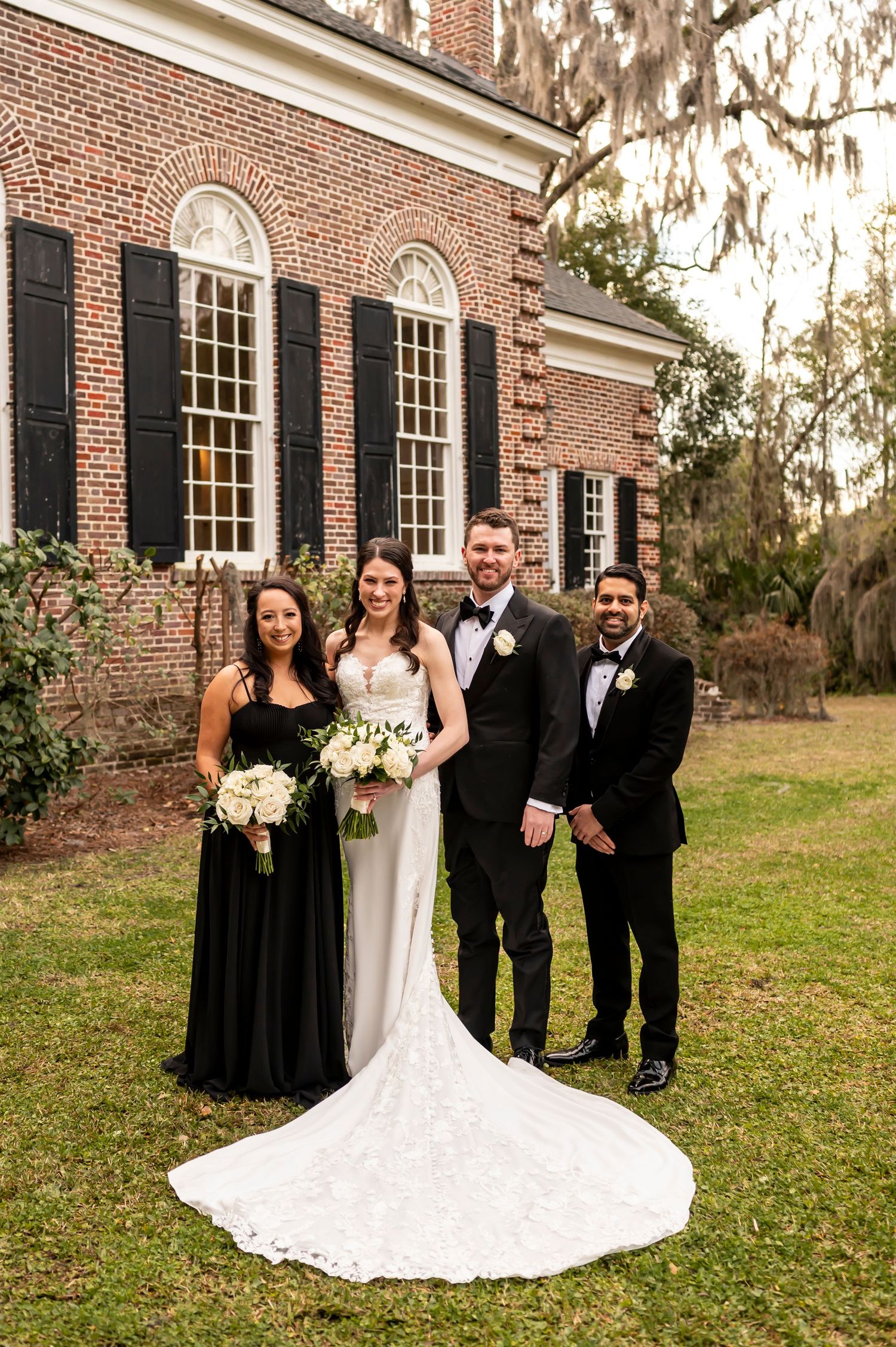 Wedding party poses in front of historic red brick colonial building with long white train on dress and black attire.