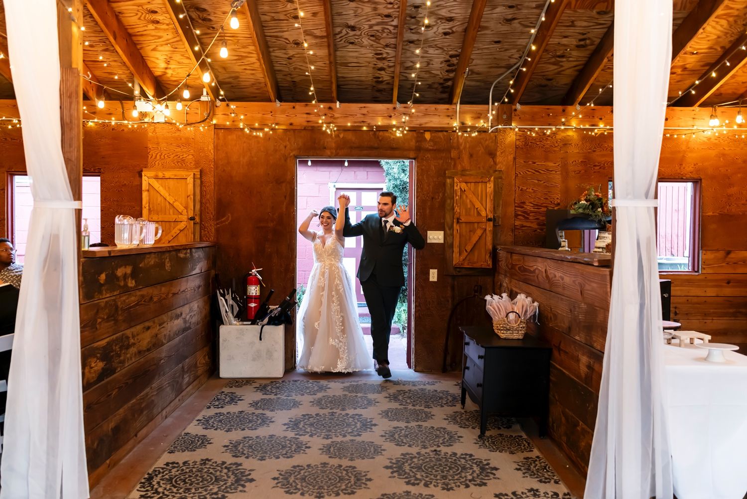 A rustic wooden barn venue decorated with string lights and white fabric drapes creates a warm ambiance for a wedding celebration.
