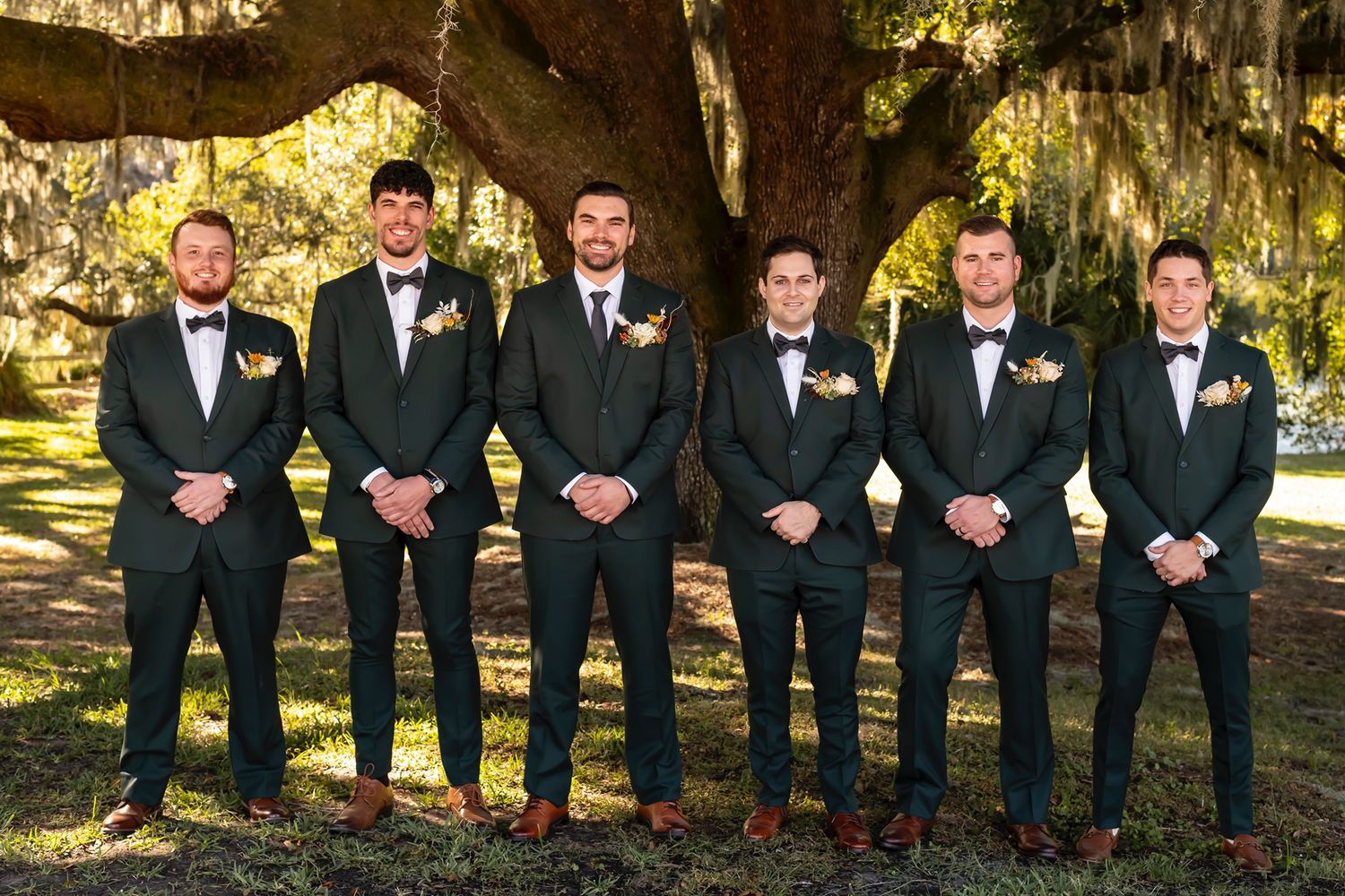Six groomsmen in formal suits and bowties pose together under a large tree draped with Spanish moss at an outdoor venue.
