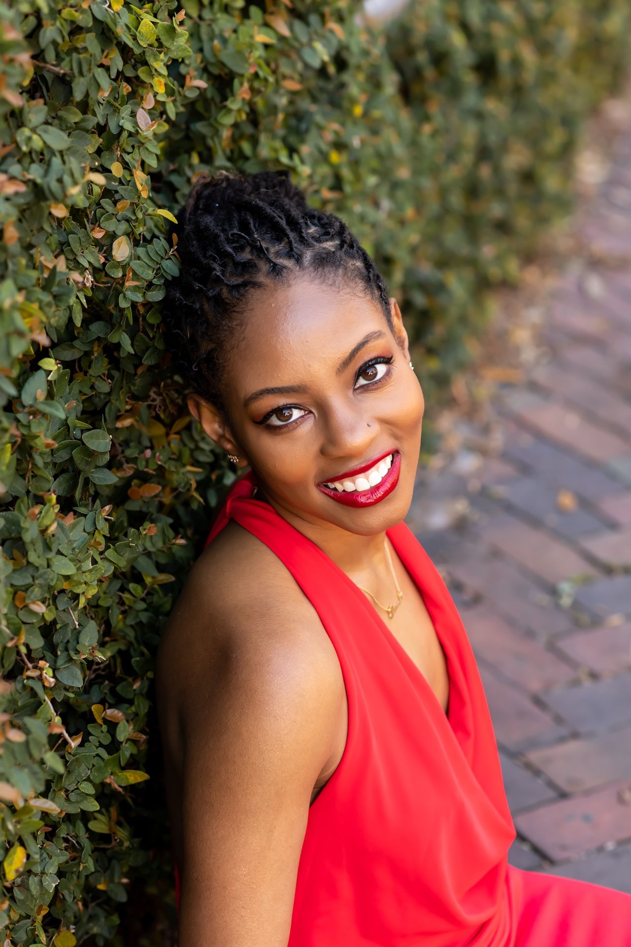 Portrait in a vibrant red sleeveless top posed against green foliage with a bright smile and braided updo hairstyle.