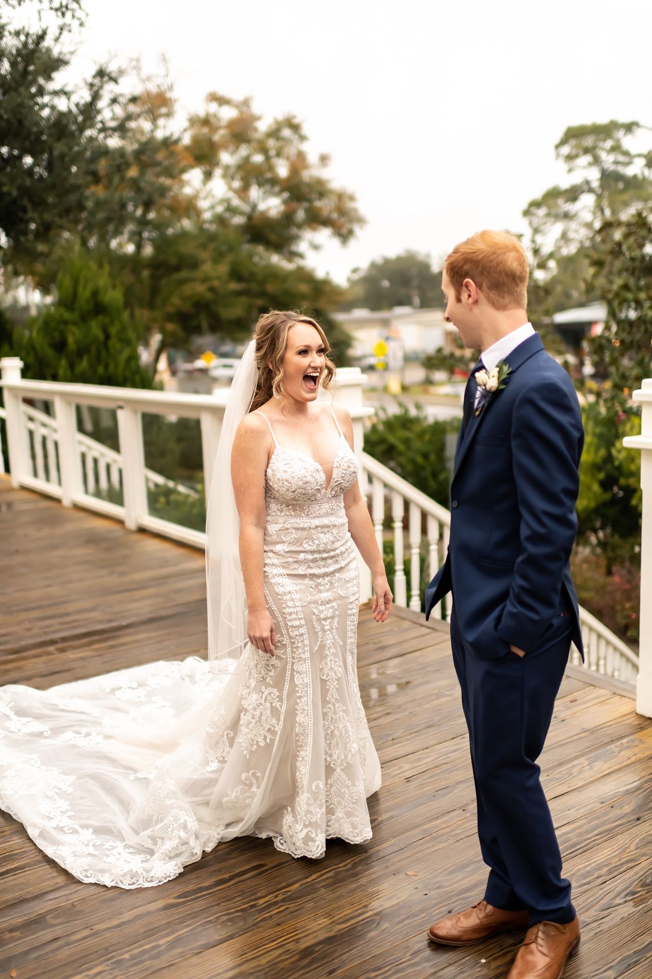 A joyful wedding couple shares a candid moment on a wooden deck with lush greenery in the background.