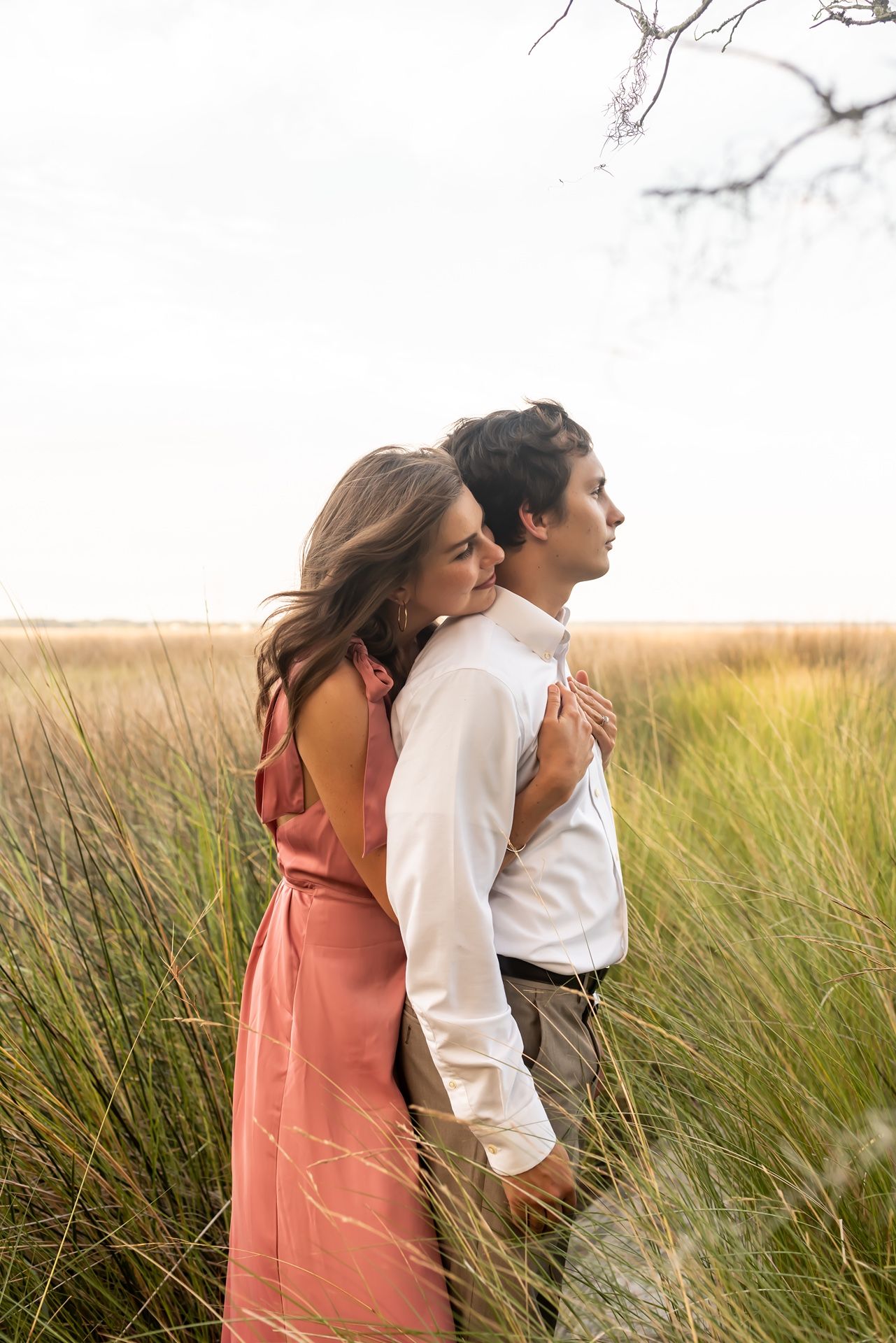 Couple embraces in golden field of tall grass during sunset as light glows warmly through their romantic moment.