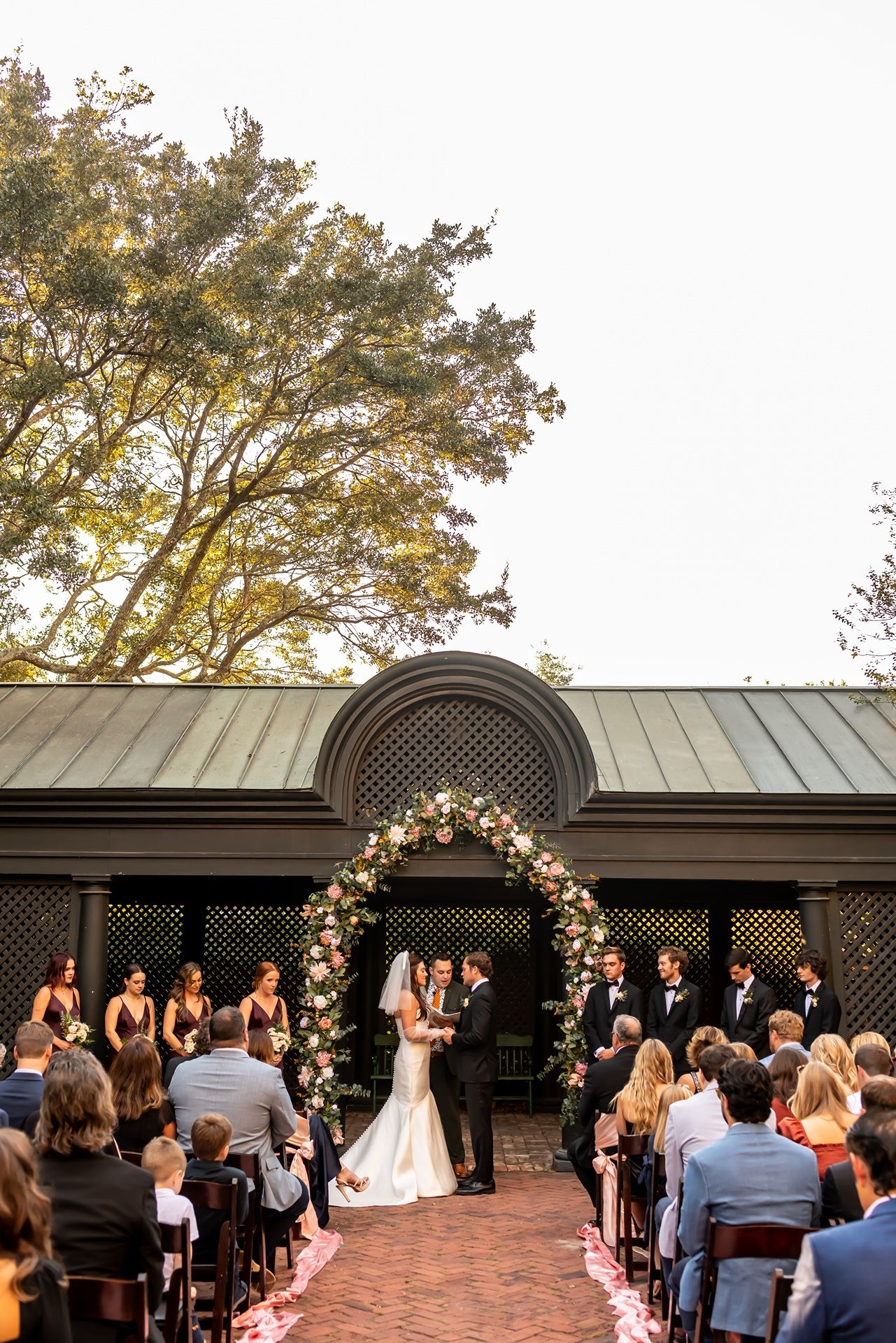 Wedding ceremony taking place under a floral arch at an outdoor venue with guests seated on chairs along a brick pathway.