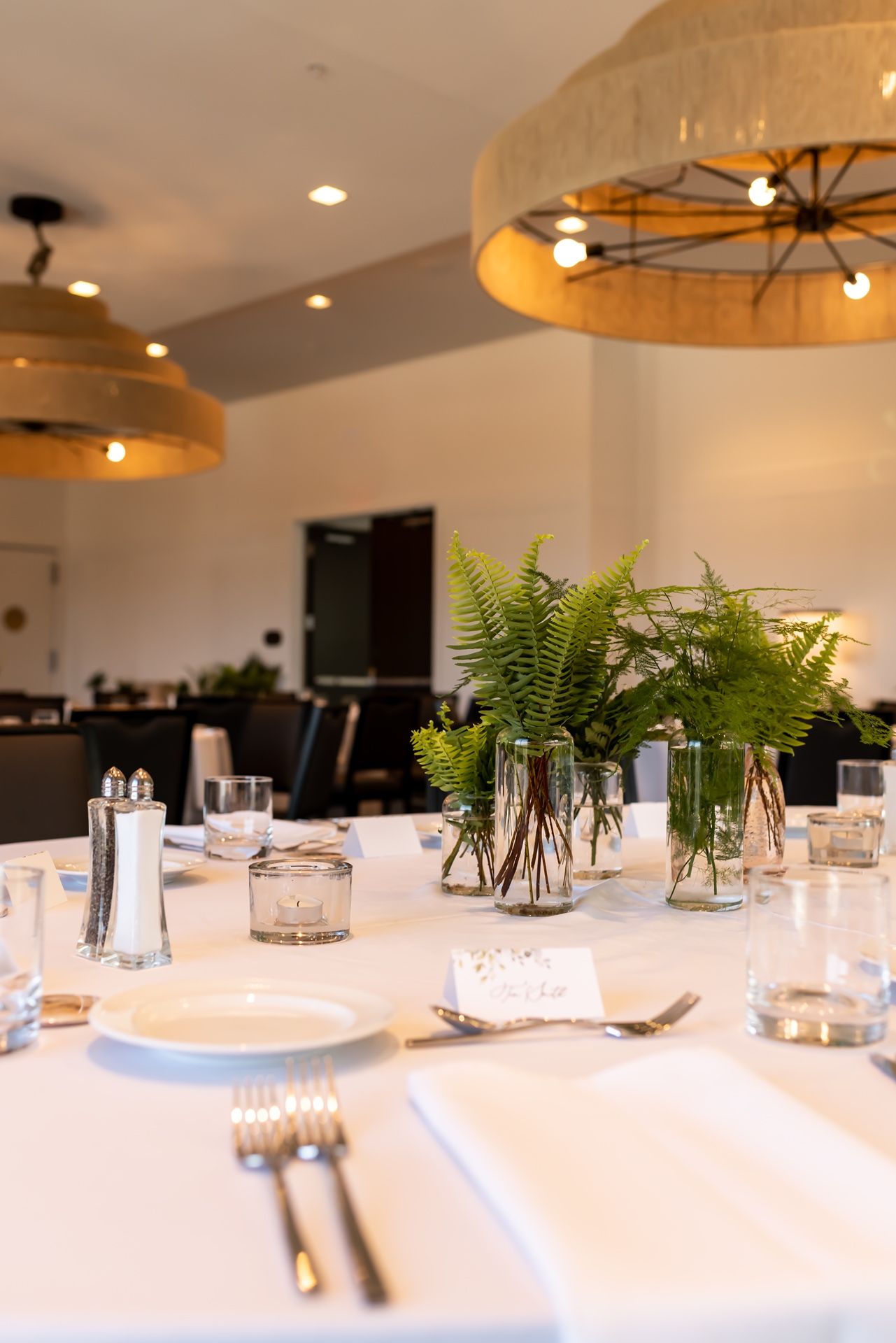 Elegant table setting with fern centerpieces in glass vases, white linens, and rustic wooden lighting fixtures above.