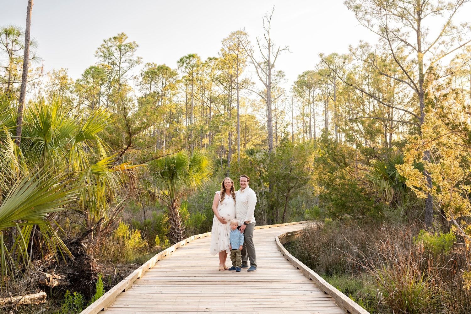 Couple in white outfits standing on a wooden boardwalk surrounded by lush green trees and palms in golden sunlight.