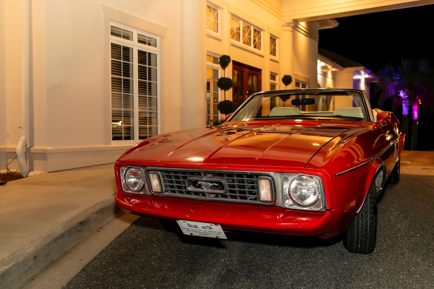 Classic red Ford Mustang parked at night outside an elegant building with white walls.