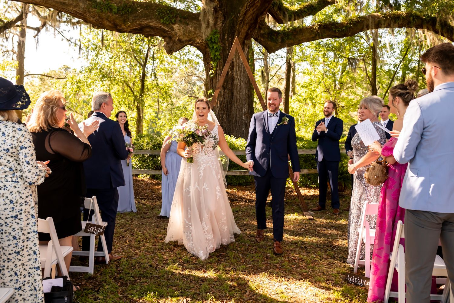 A couple walks down an outdoor aisle lined with white chairs during an autumn wedding ceremony in a garden setting.