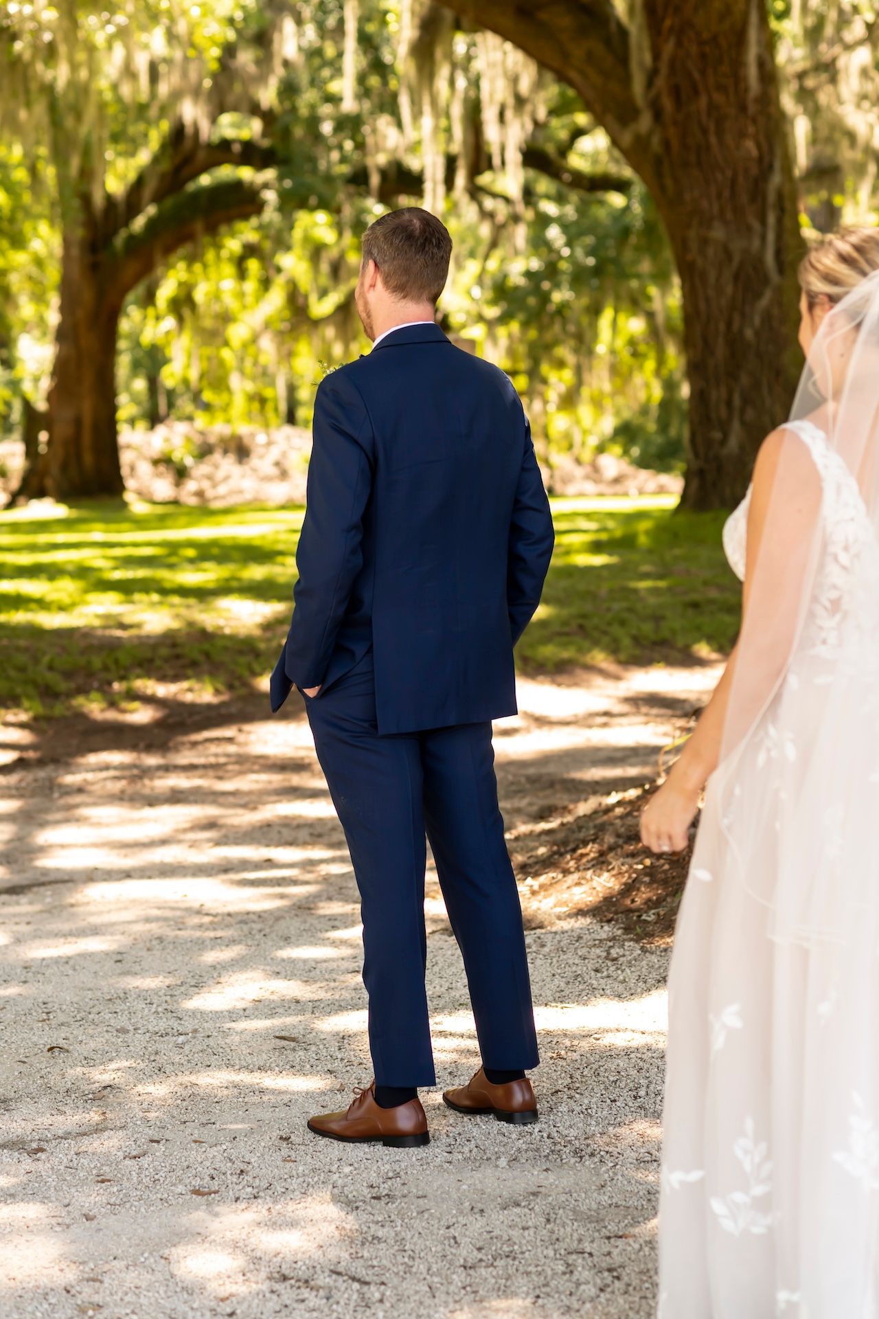 A romantic first look moment between newlyweds on a sunlit path under oak trees draped with Spanish moss.