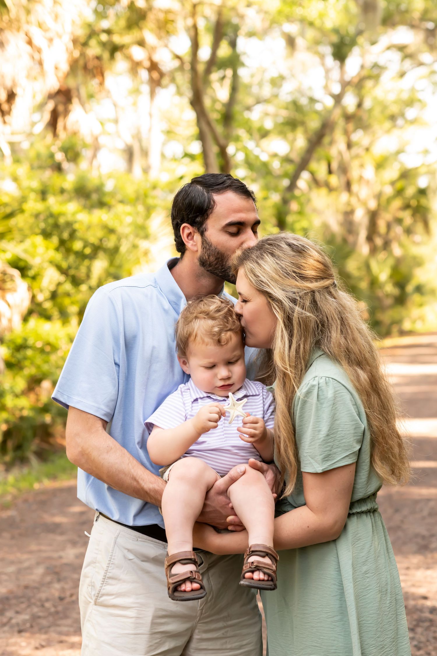A couple share tender moments with their baby while standing on a tree-lined path during golden hour.