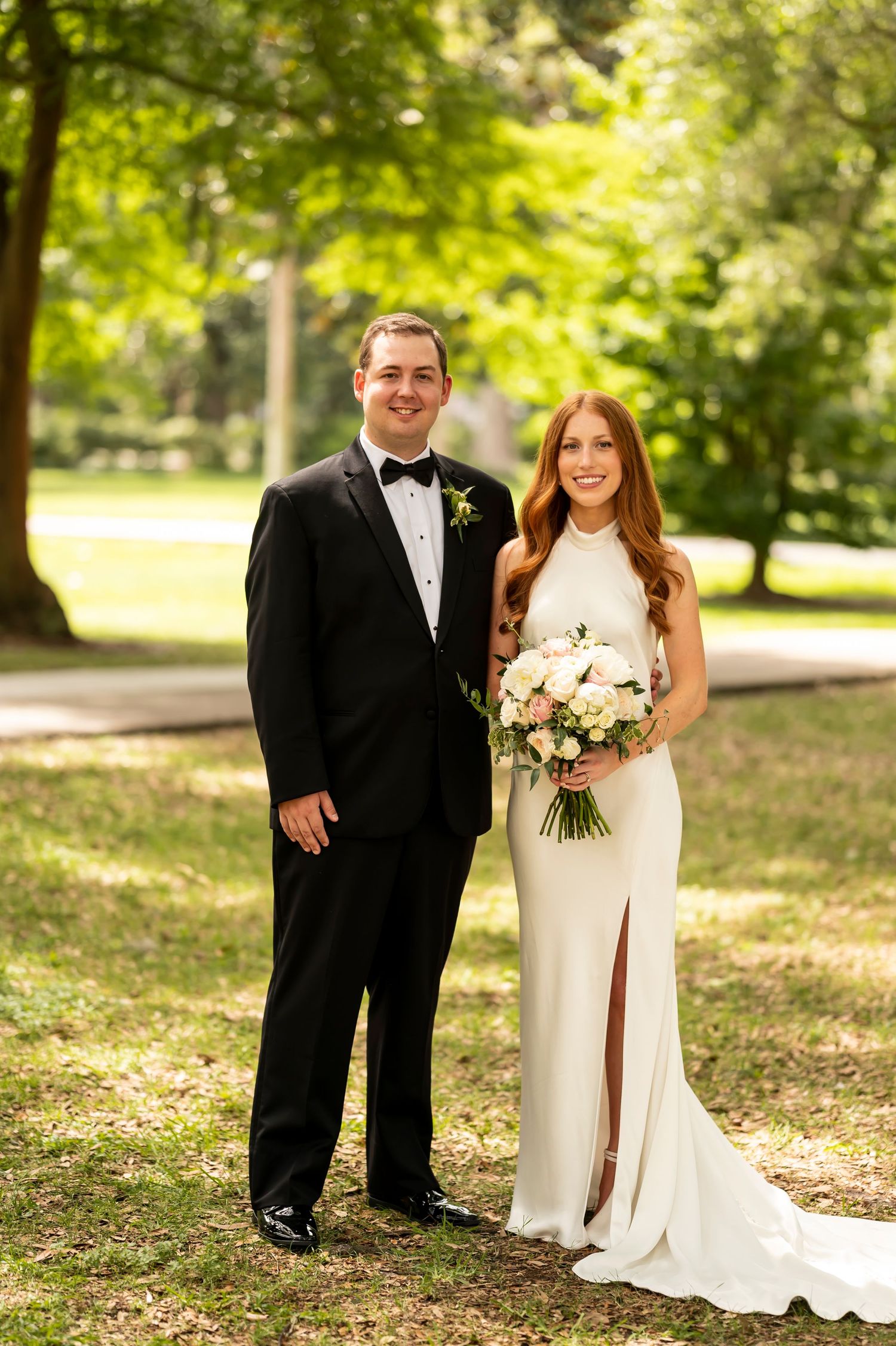 A couple stands together outdoors in wedding attire with the bride holding a white floral bouquet in a sunlit wooded setting.