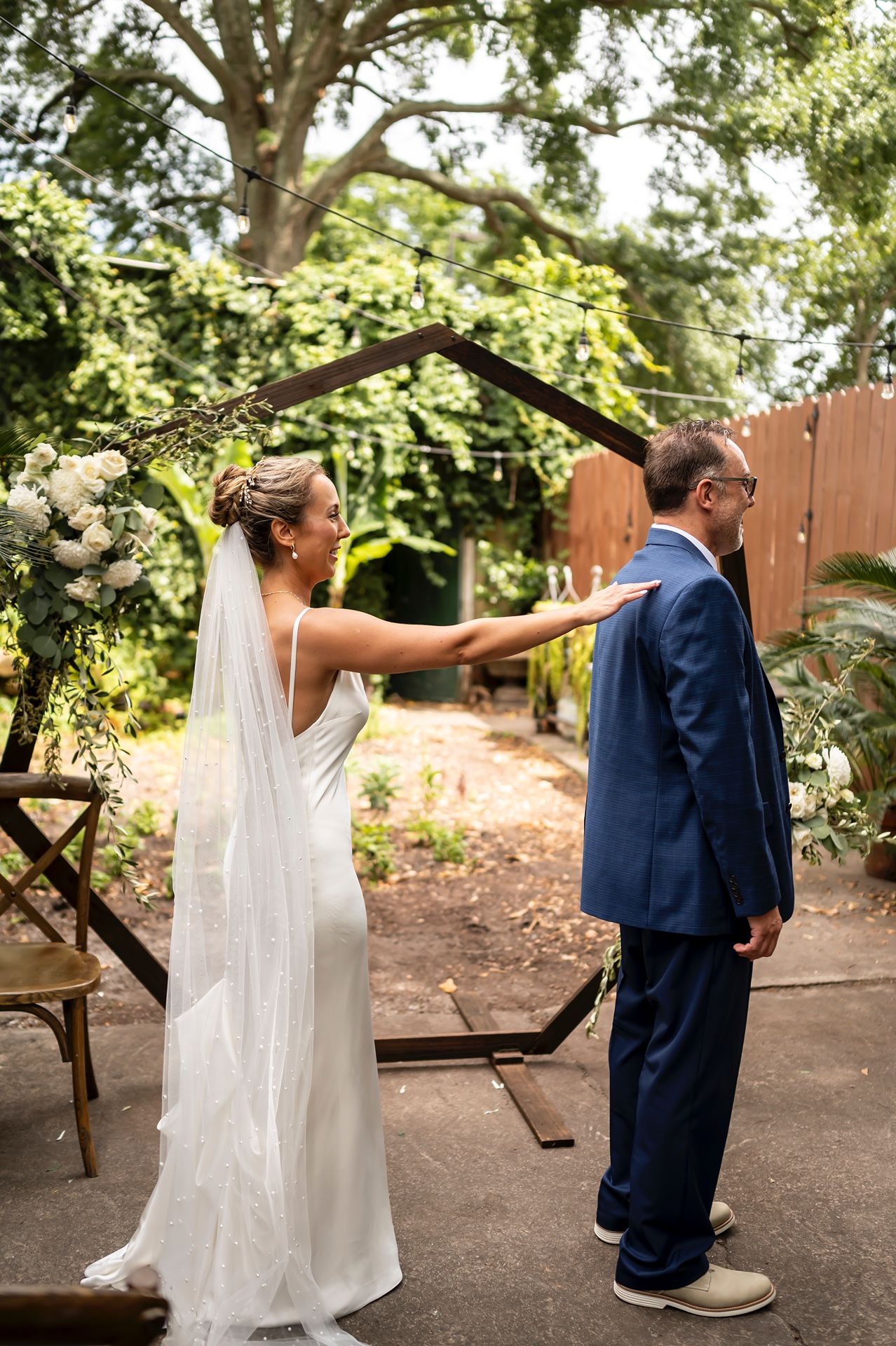 Wedding couple plays a game during their outdoor garden ceremony with bride tapping groom's shoulder playfully.