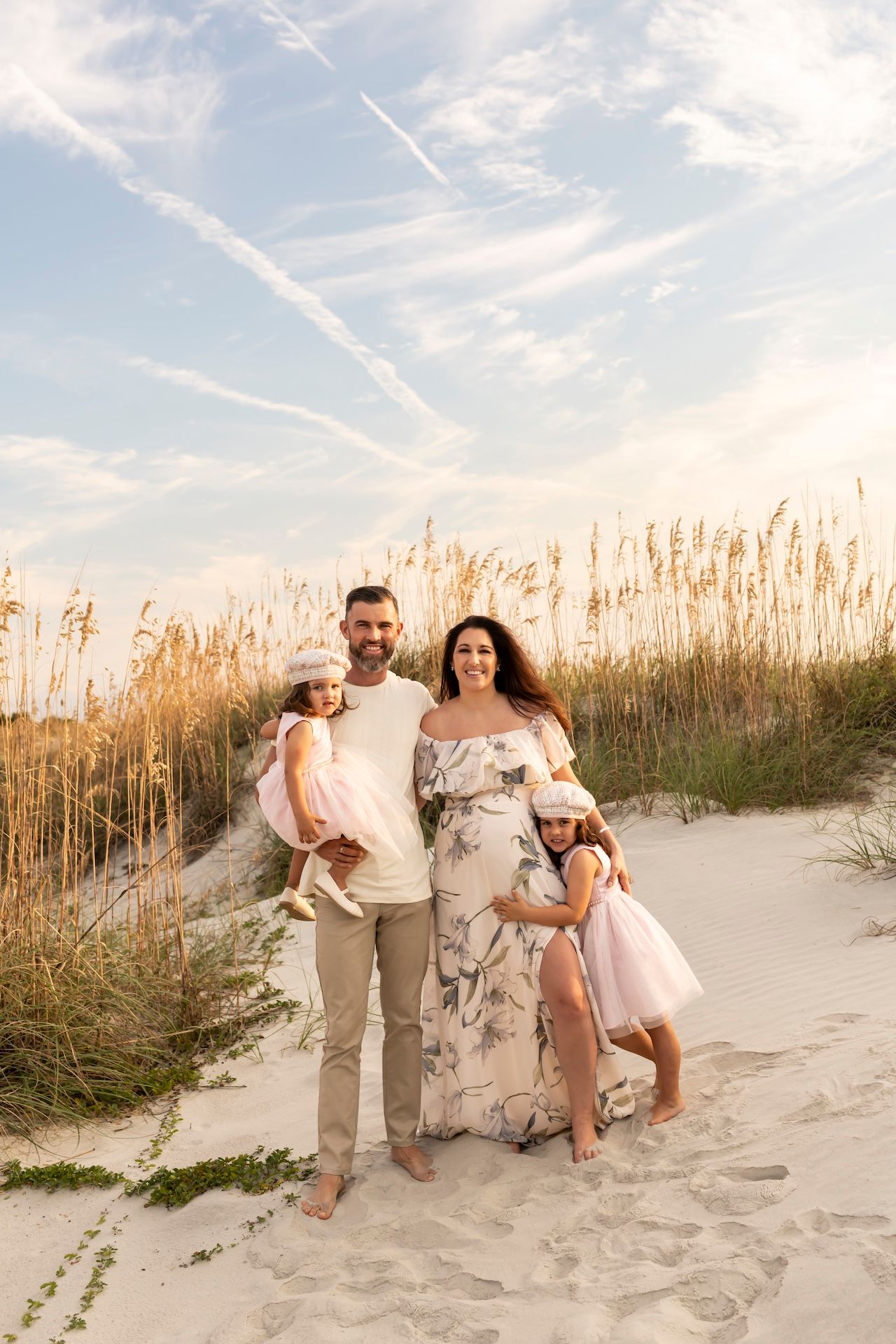A family enjoys a sunset moment together on sandy dunes with tall beach grass and wispy clouds in the sky.