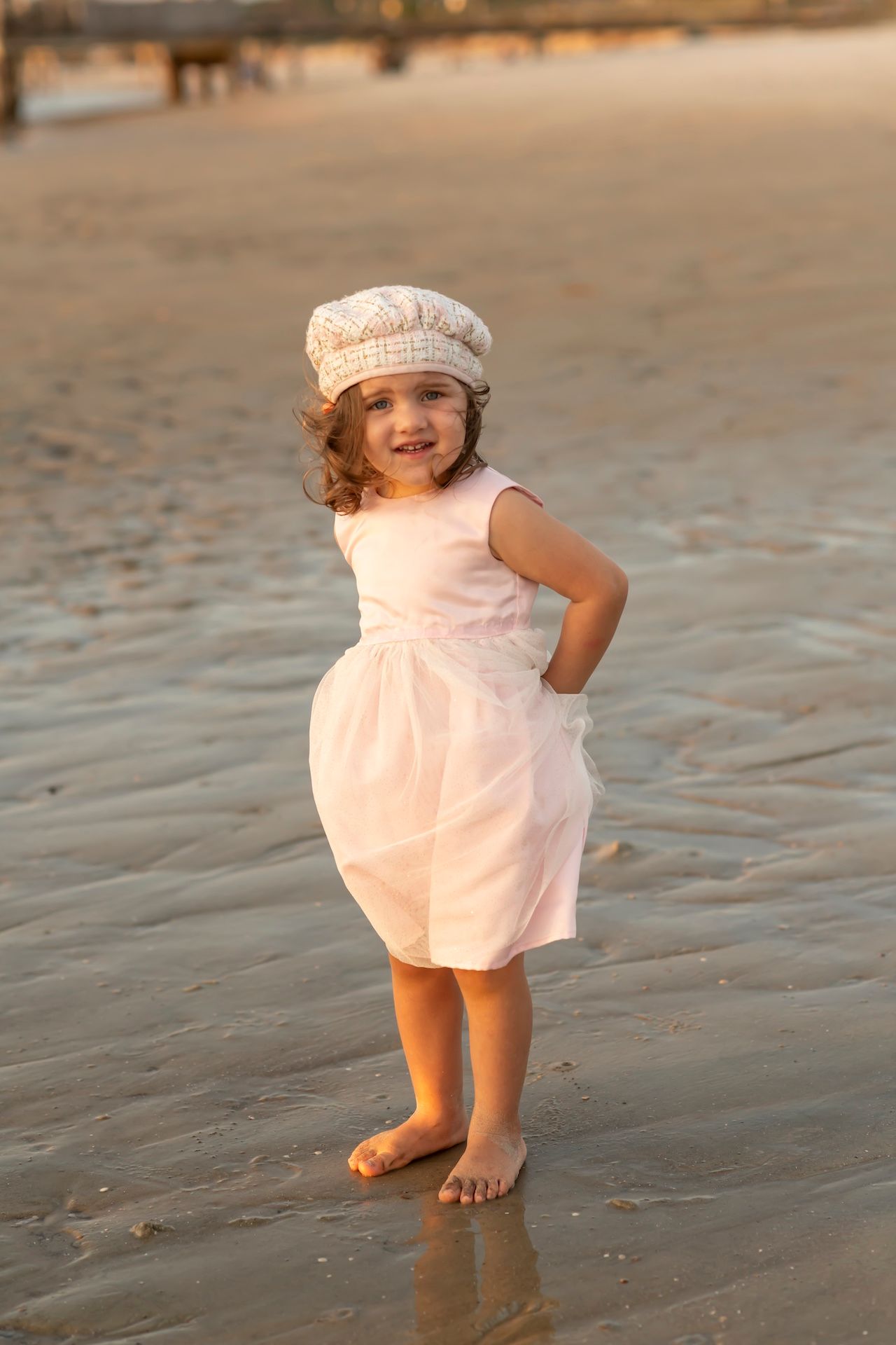 A young child wearing a pink dress and knit hat stands barefoot on a beach at sunset.