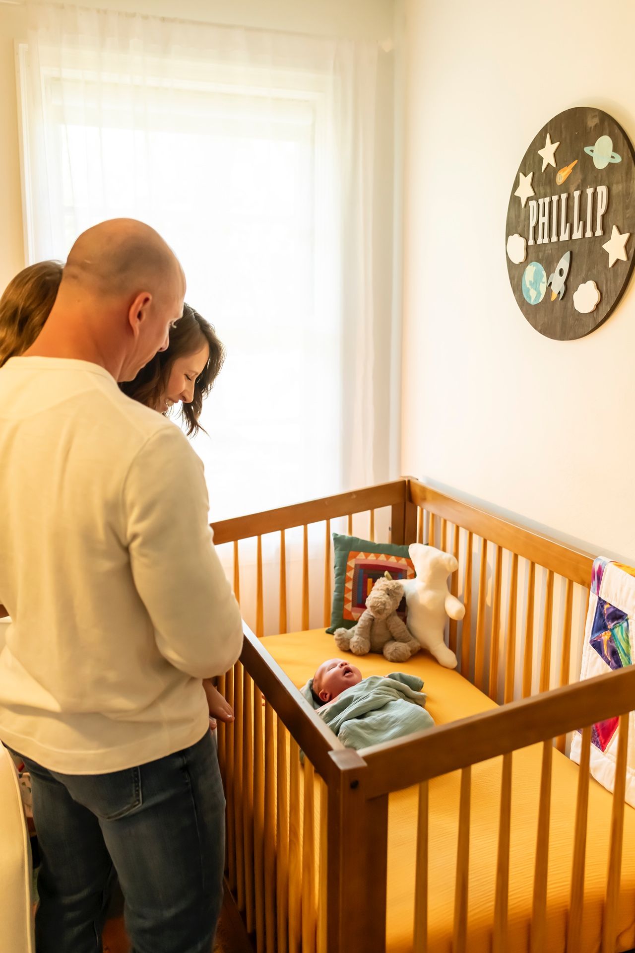 Two people standing by a wooden crib with a circular sign reading 'Phillip' decorated with stars on the nursery wall.