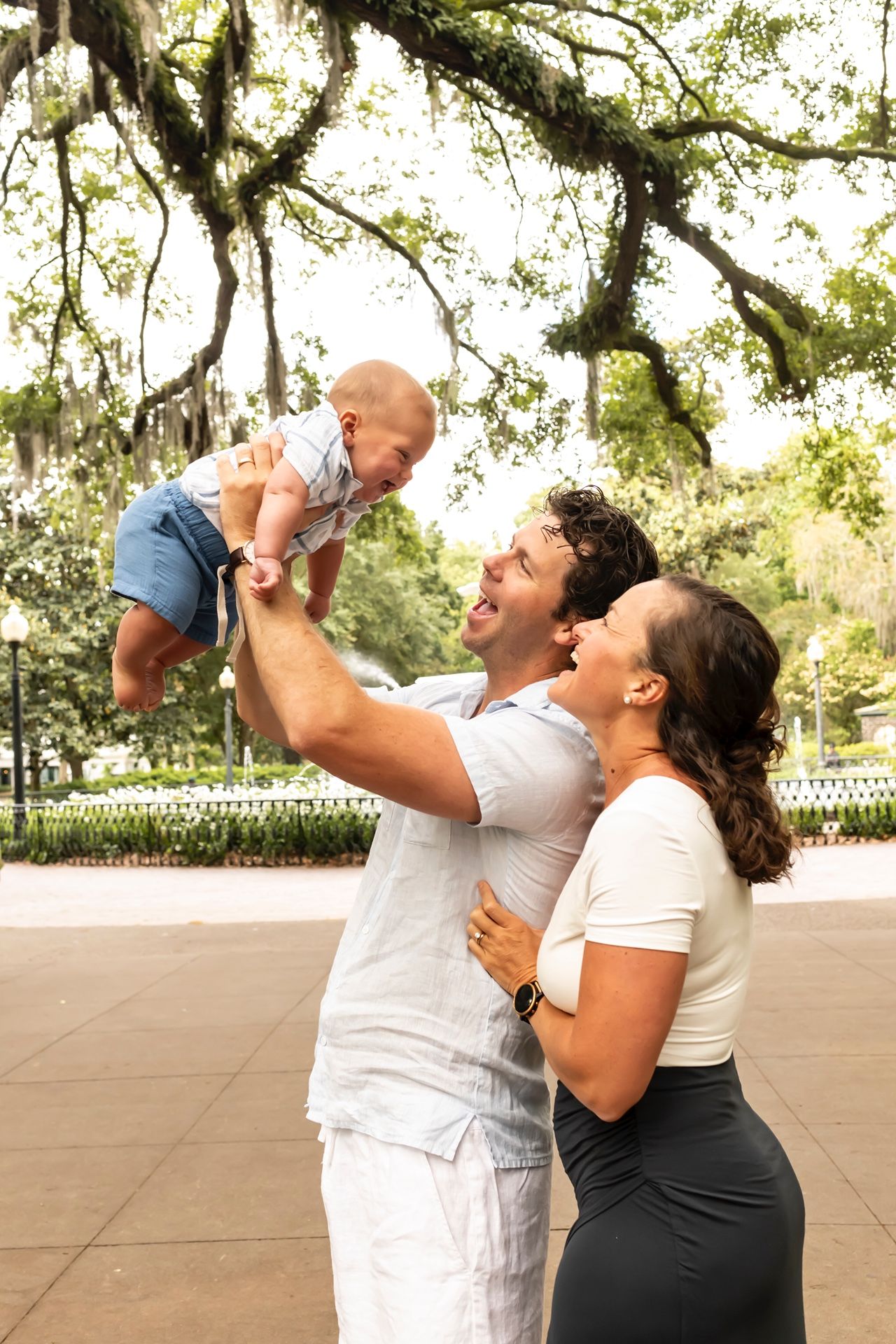 A delighted family plays together as they lift their baby in the air under moss-covered trees.
