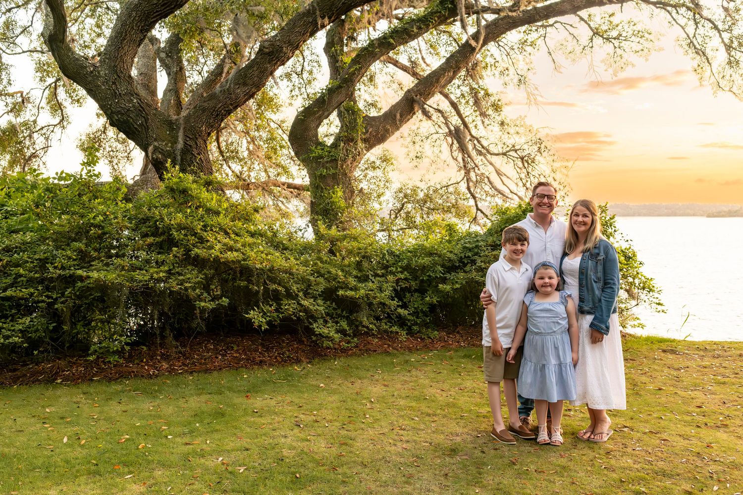 A family enjoys a sunset moment together on a grassy lawn beneath gnarled trees near the waterfront.