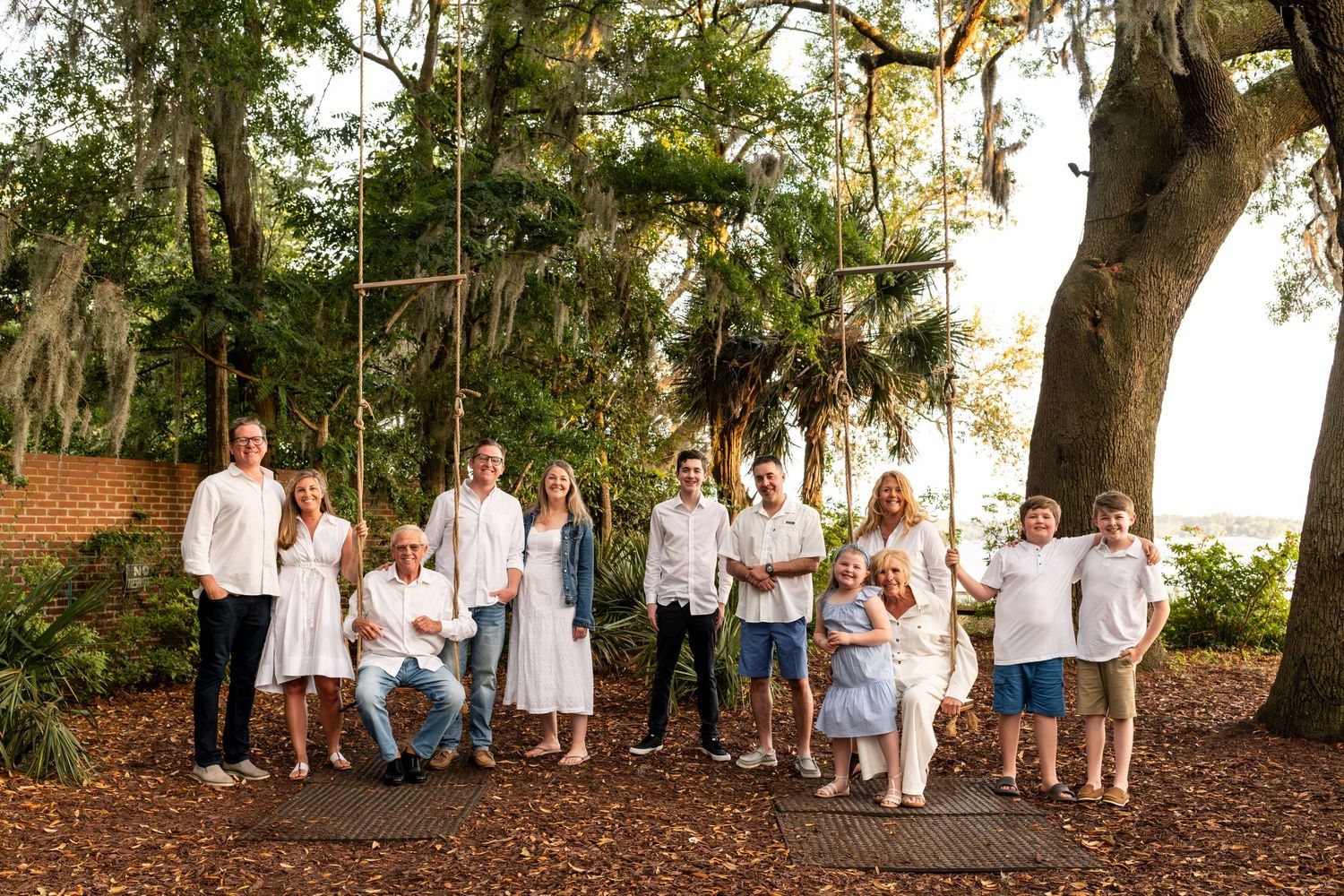 Group in white outfits poses for candid photos under large oak trees at sunset.
