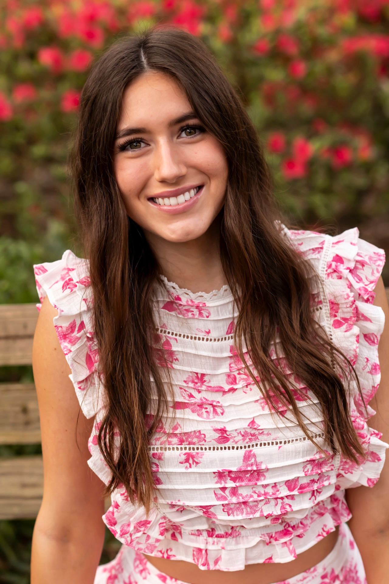 Person wearing a white and pink floral ruffle top poses in front of blooming red flowers outdoors.