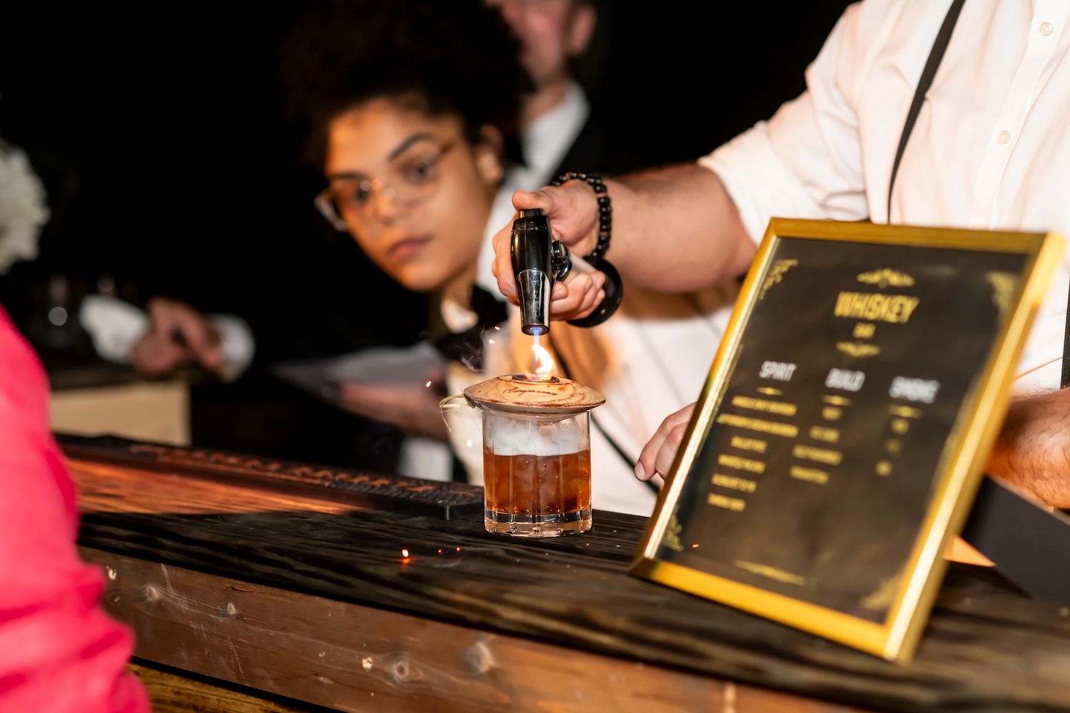 Bartender pouring a craft cocktail at a dimly lit bar with golden menu board in view.