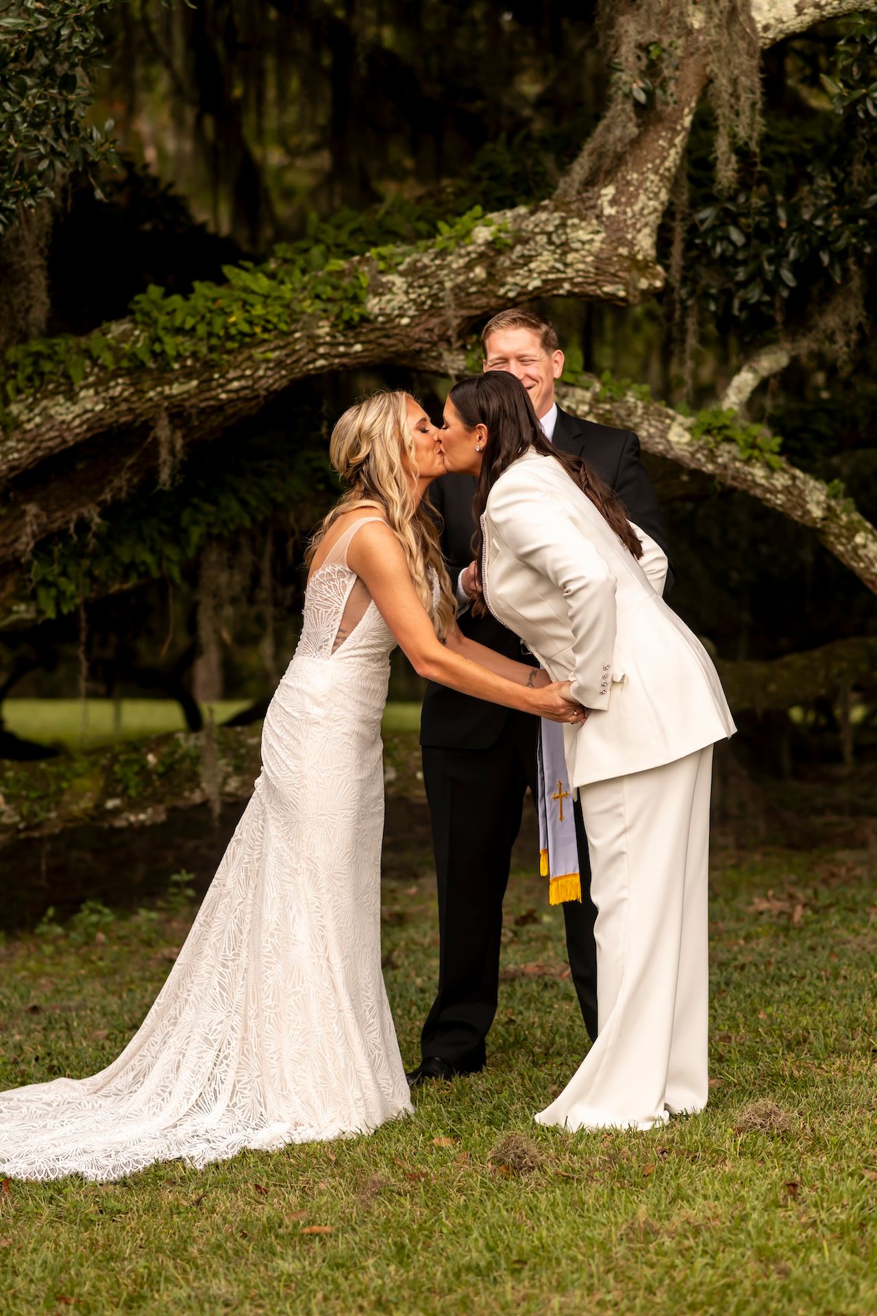 Two people in white wedding attire share a tender moment on a grassy lawn beneath moss-draped trees at sunset.