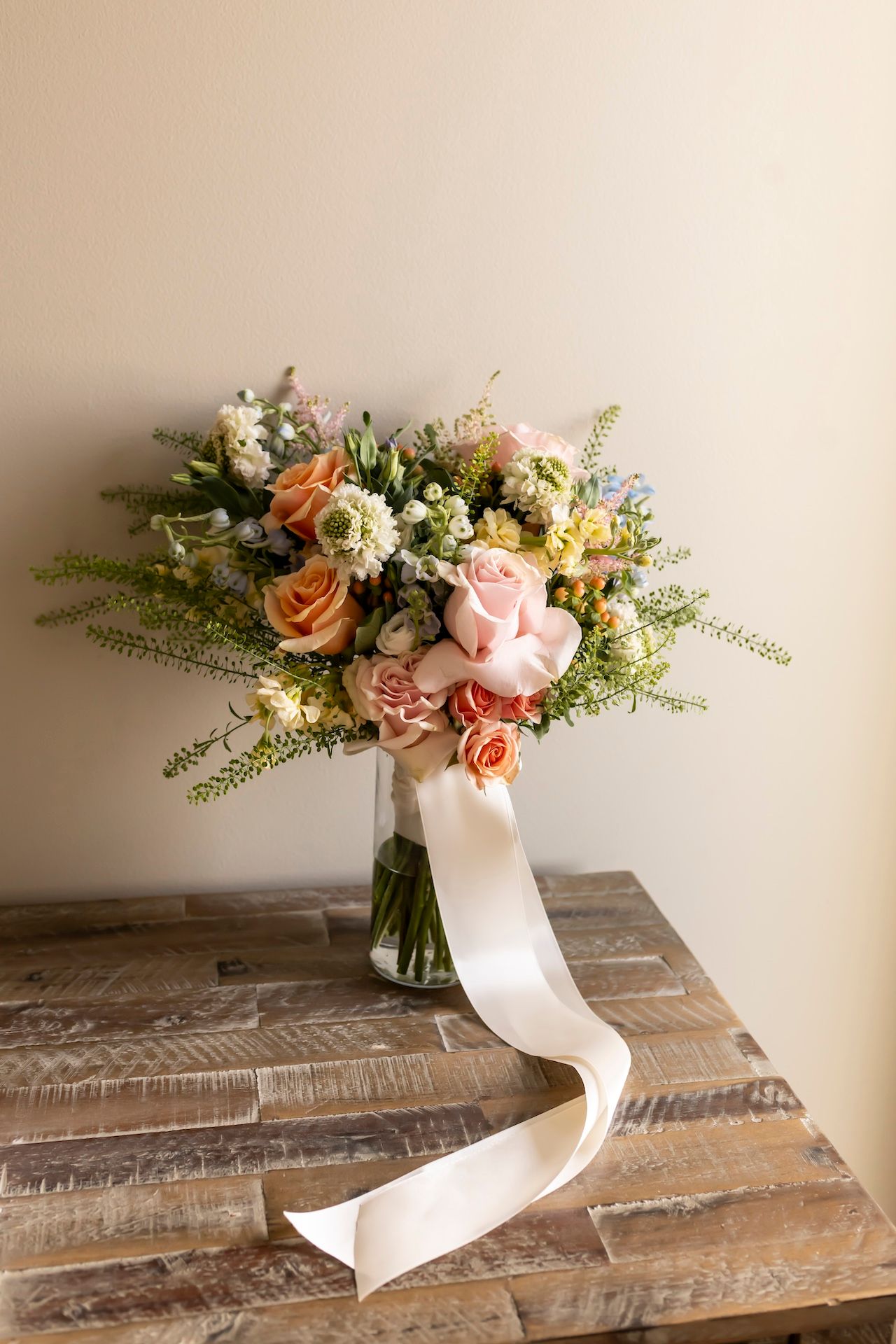 Romantic bridal bouquet with pink and peach roses tied with flowing white ribbon sits on rustic wooden table.