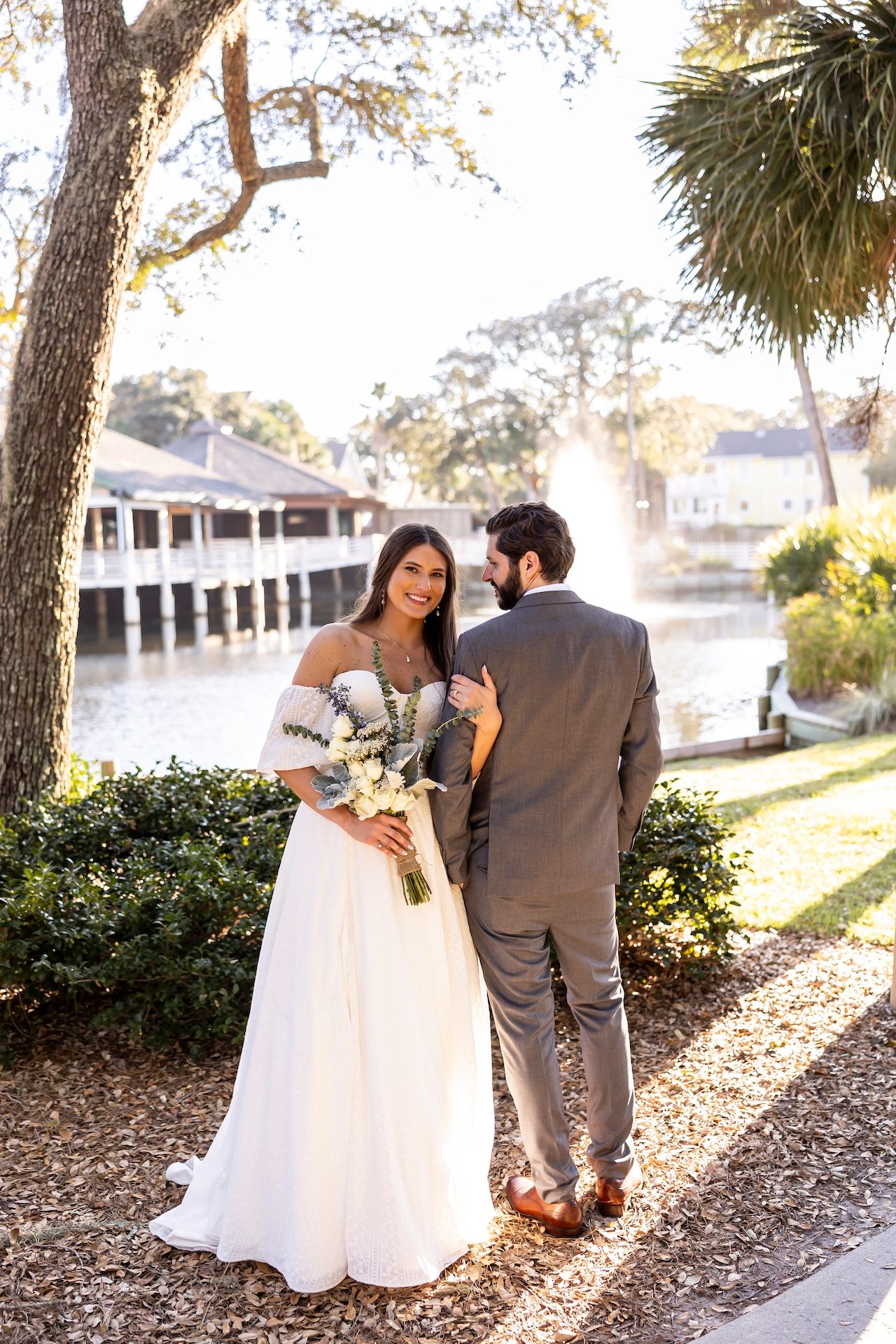 Newlywed couple standing beside water at outdoor venue with white dress and grey suit among trees and greenery.