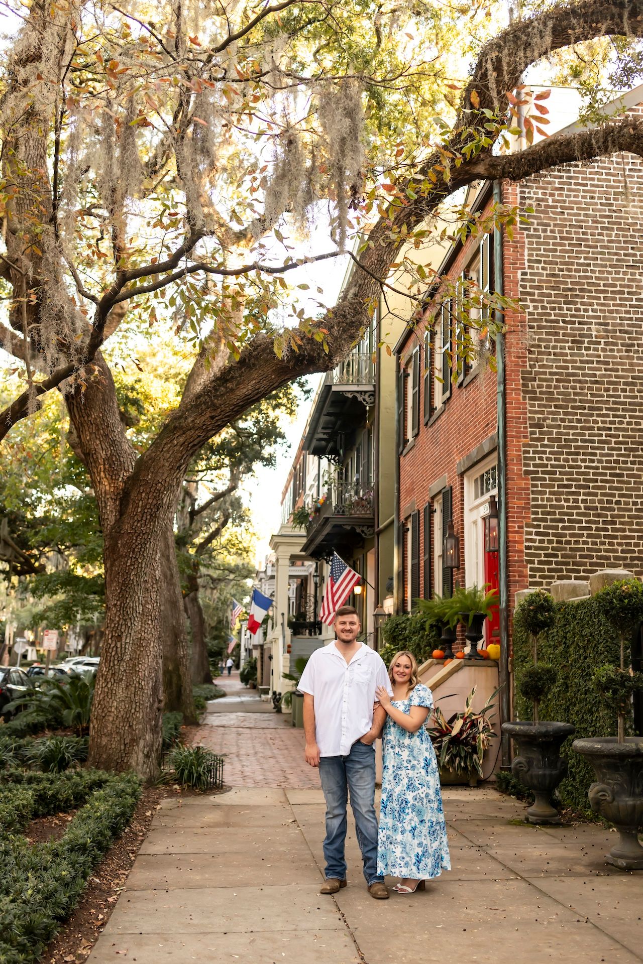 A couple poses on a historic brick-lined street with Spanish moss hanging from trees above.