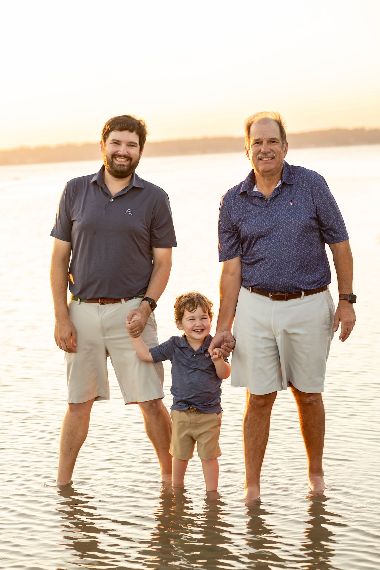 A family enjoying a sunset walk in shallow water at the beach, all wearing matching navy shirts and khaki shorts.