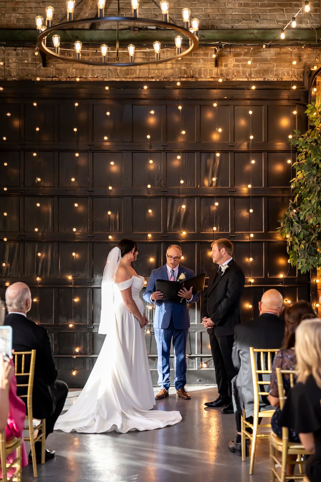 A wedding ceremony takes place in an intimate venue with exposed brick walls and romantic string lights overhead.