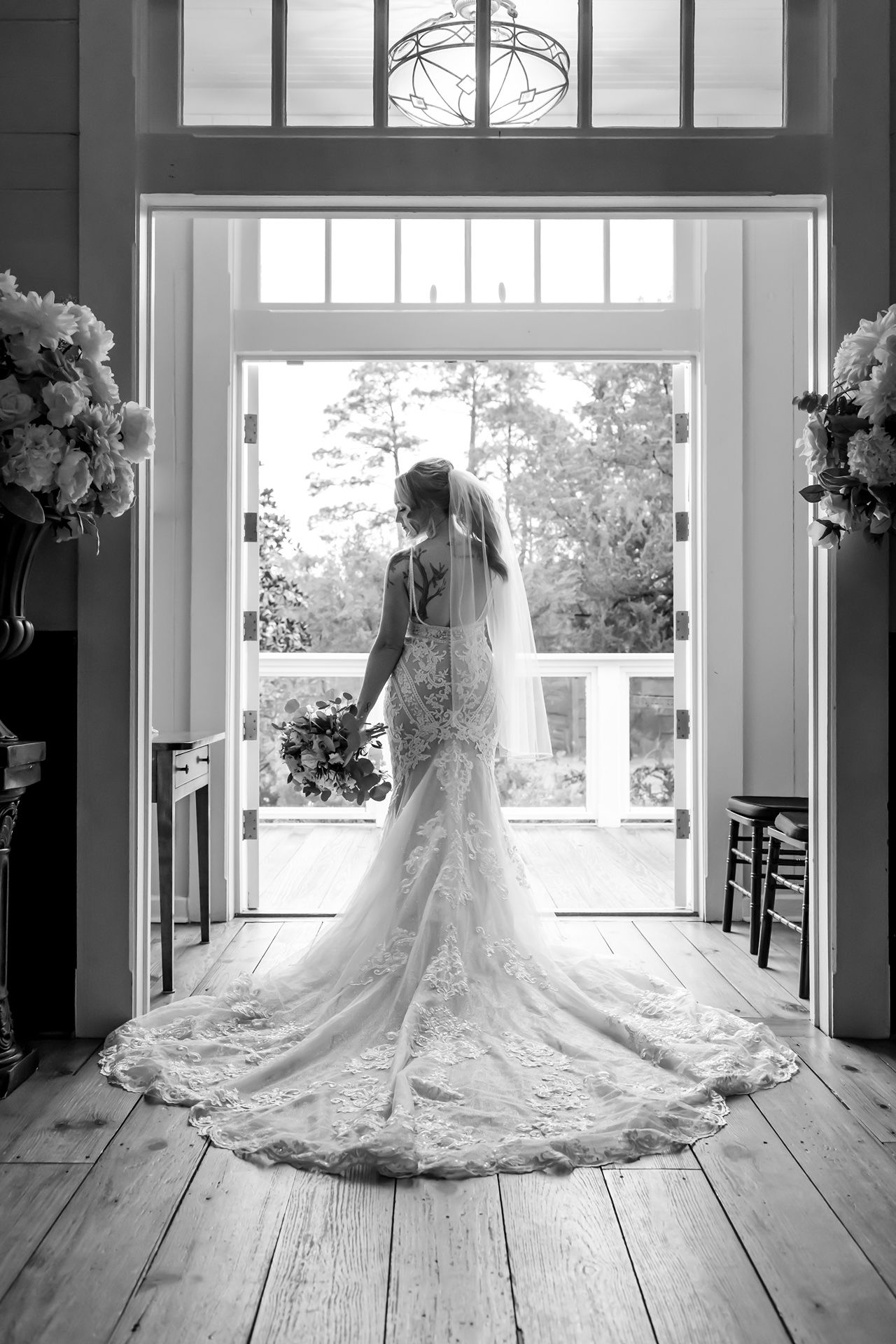 Dramatic bridal portrait in black and white shows flowing lace dress and veil against bright windows.