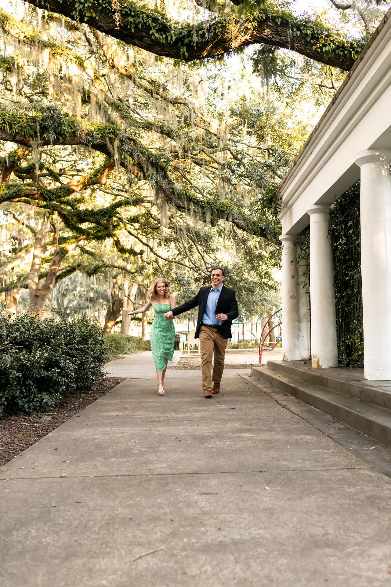 A sequence of people walking under a covered colonnade with Spanish moss hanging from trees overhead.