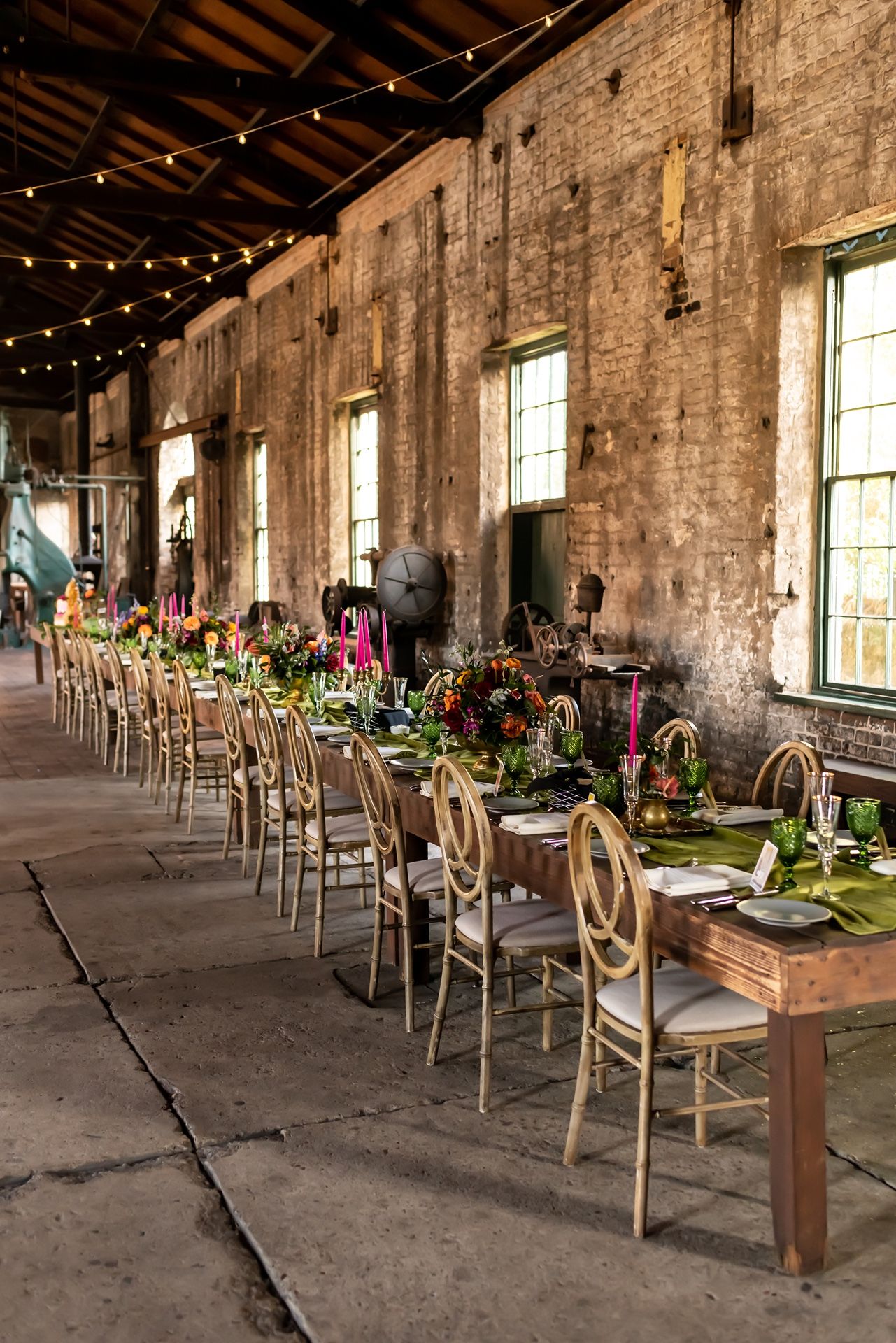 Long rustic barn venue with exposed brick walls, string lights, and wooden farm tables decorated with floral arrangements.