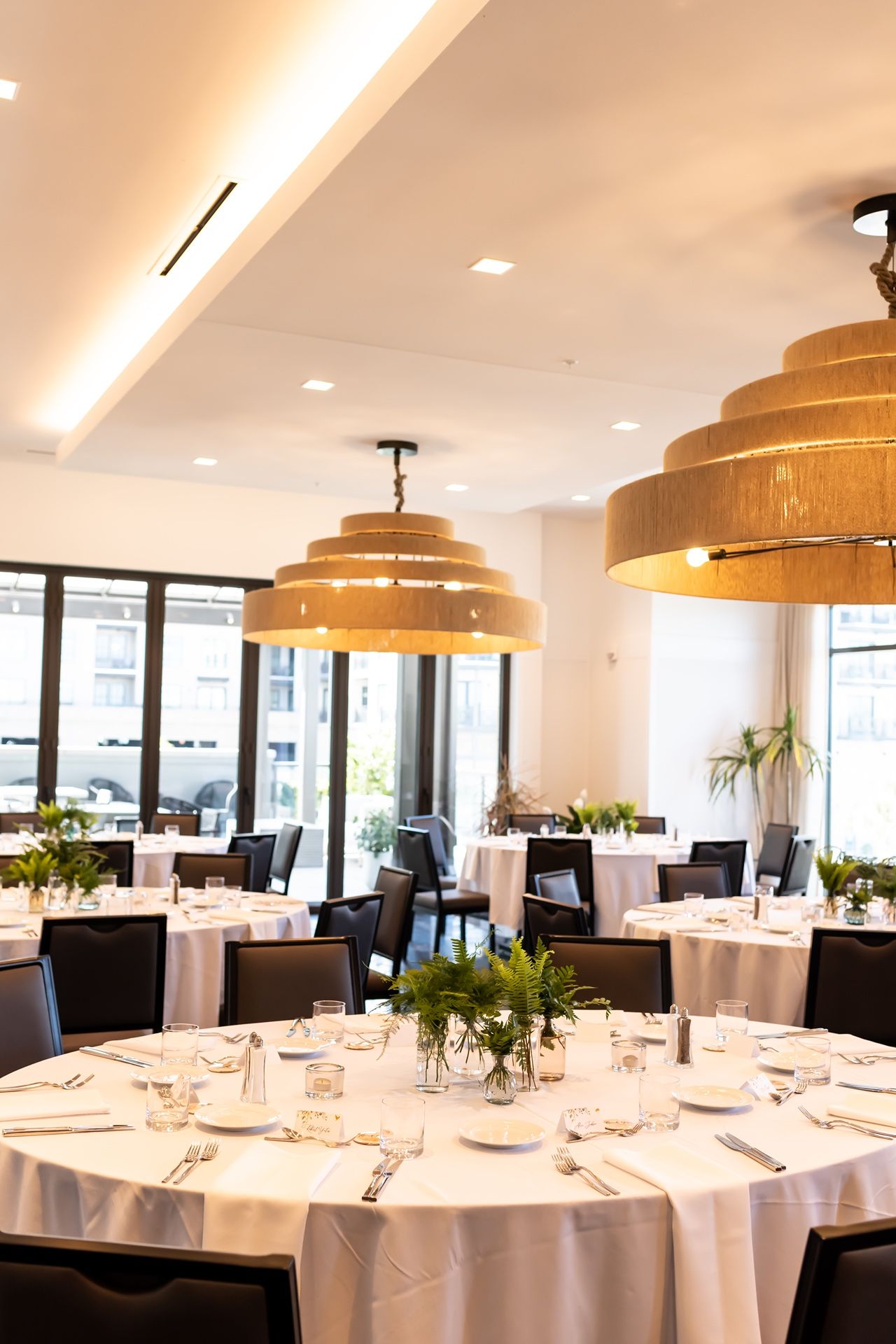 Elegant dining room with round tables set with white linens under large wooden tiered pendant lights by bright windows.