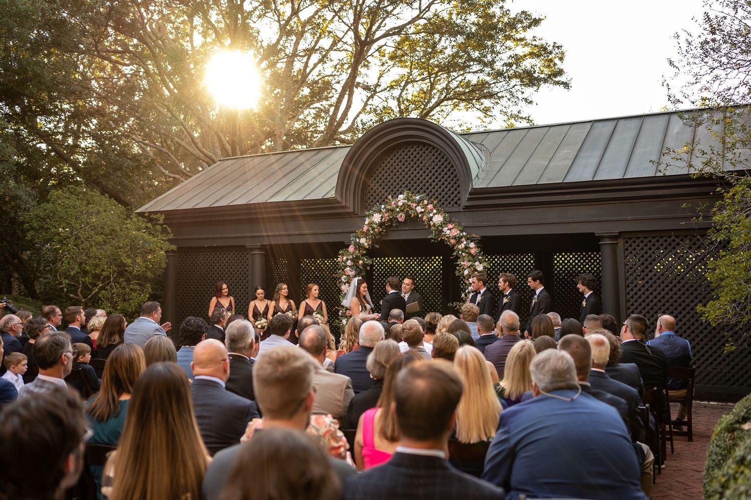 A crowd gathers for an outdoor wedding ceremony at a garden venue during golden hour sunset.