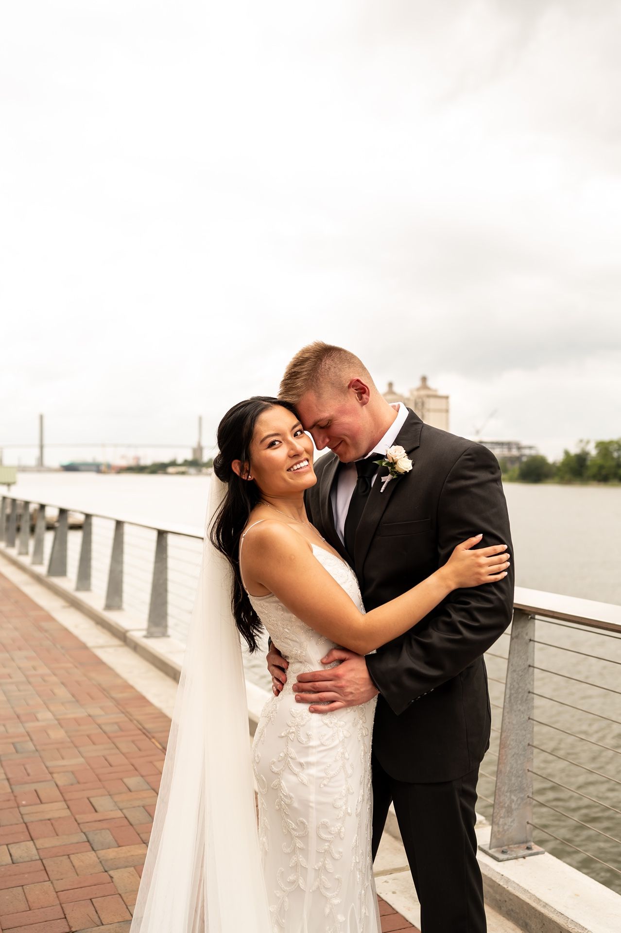A romantic waterfront wedding moment featuring a couple in formal attire embracing by a brick walkway.