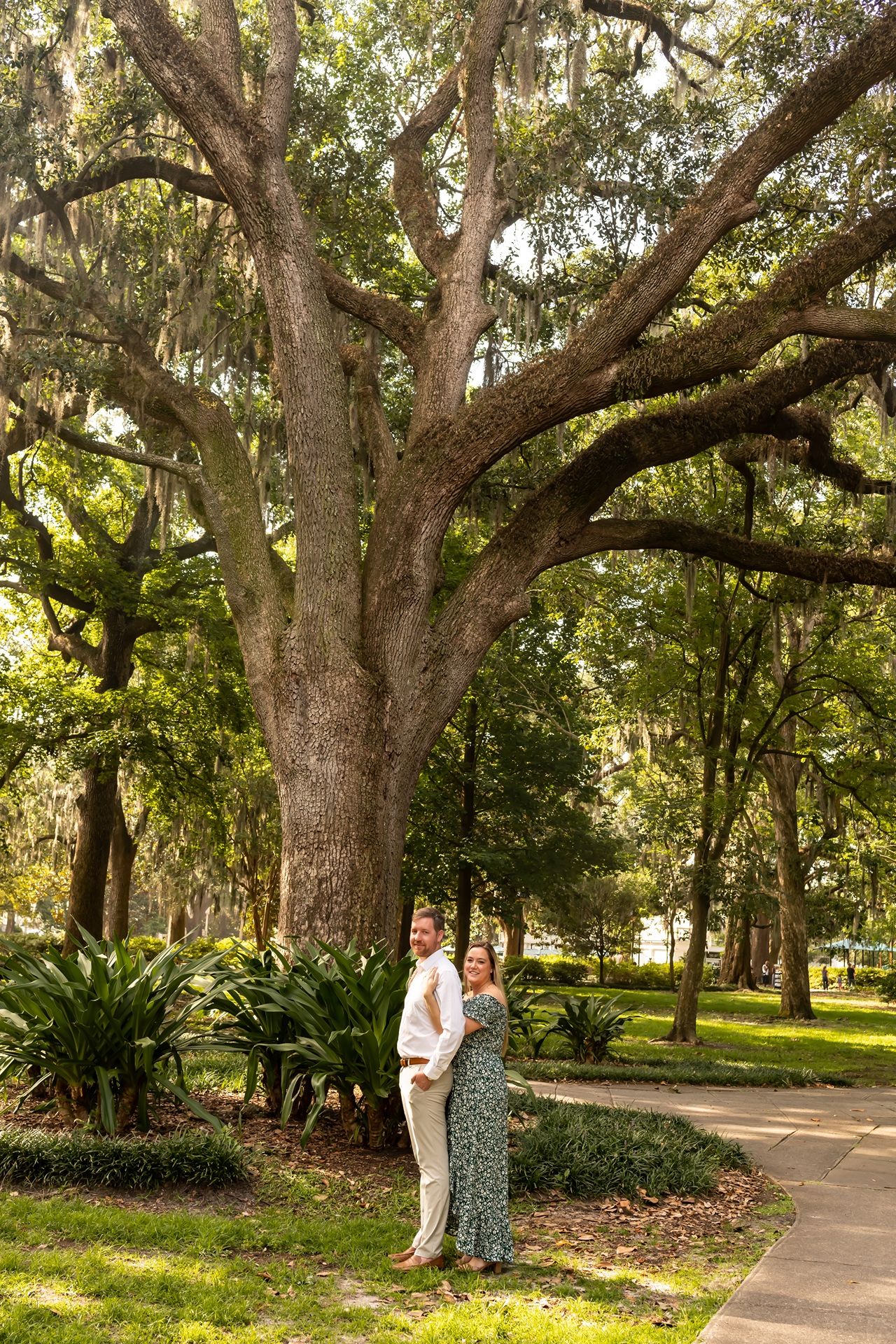 A romantic couple stands beneath a majestic live oak tree in a serene park with Spanish moss and lush landscaping.