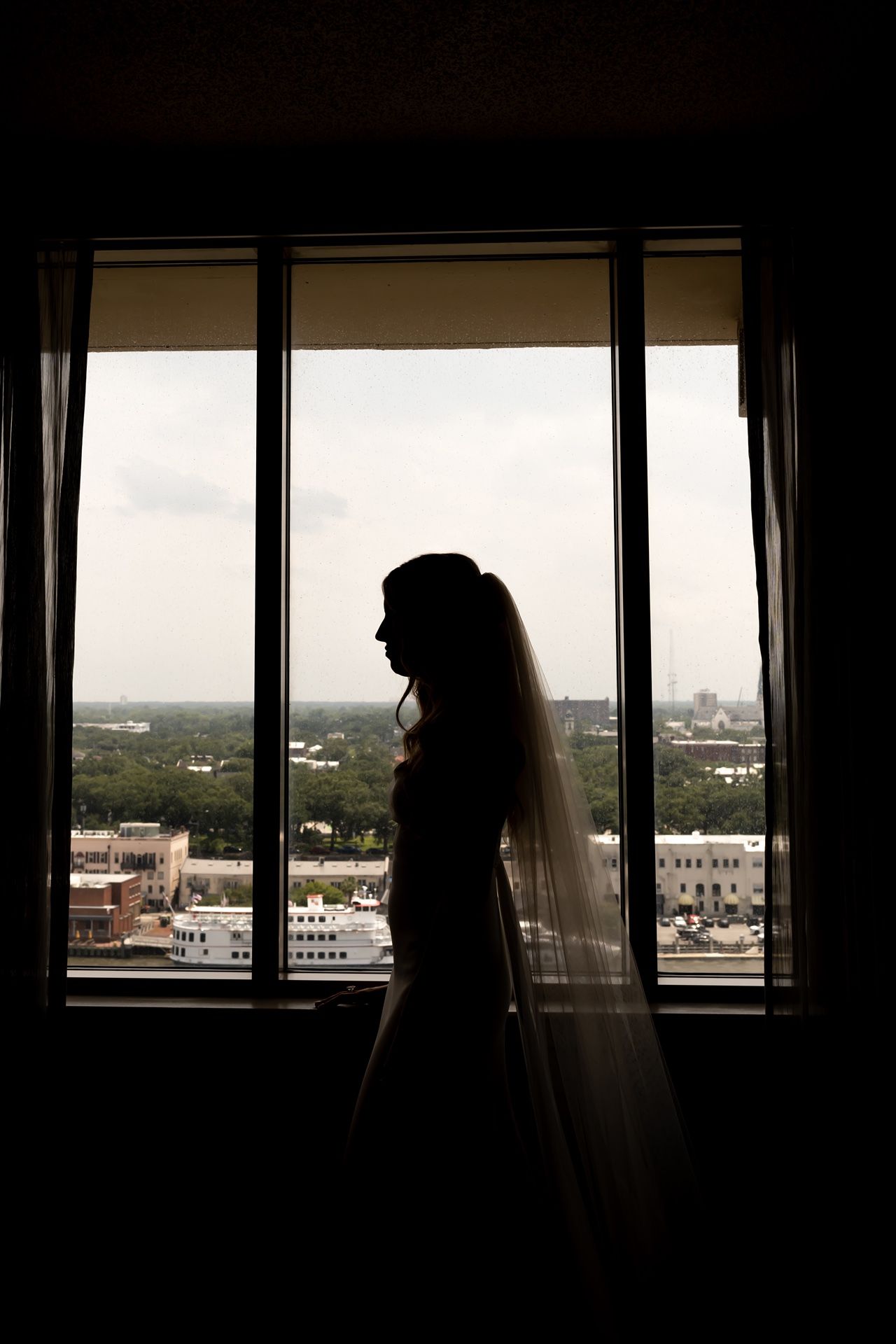 Silhouetted figure in wedding veil stands by large window overlooking cityscape view at sunset.