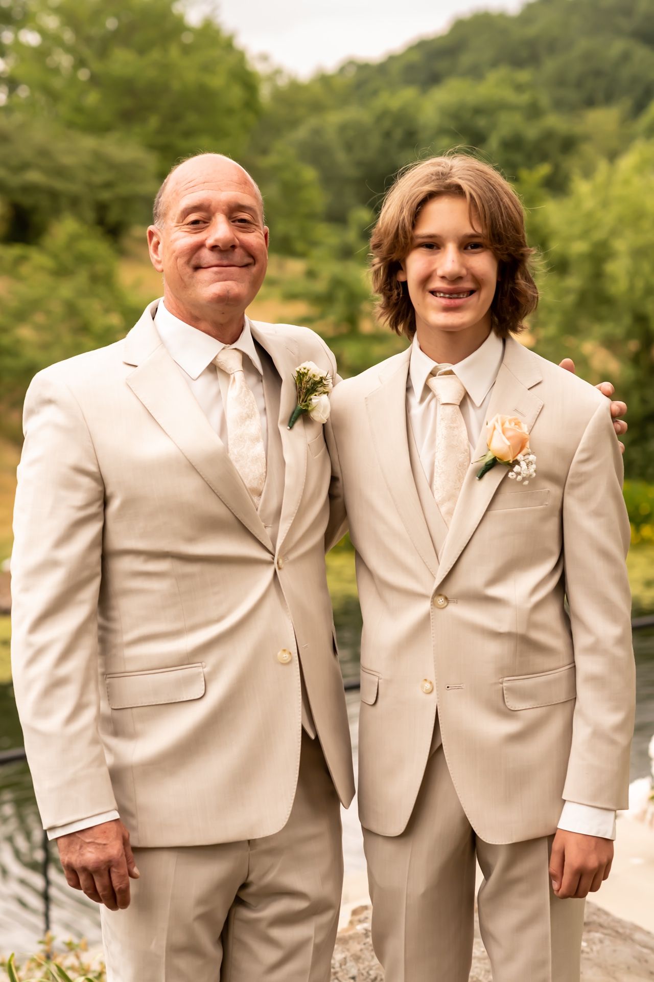 Two people in matching beige suits and ties stand together outdoors near a lake with greenery in the background.