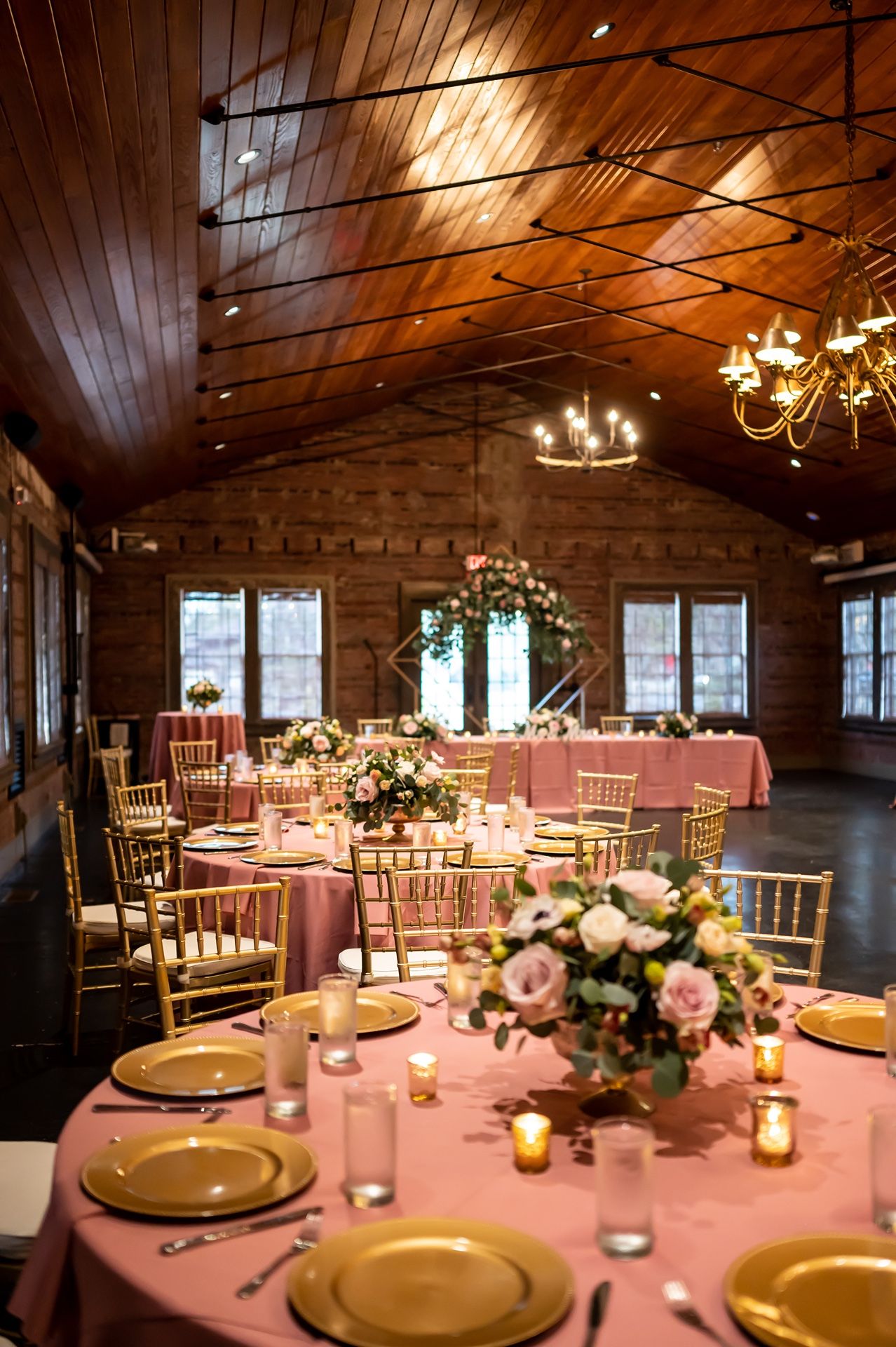 Rustic barn wedding reception venue with wooden ceiling, chandeliers, and pink floral centerpieces on round tables.