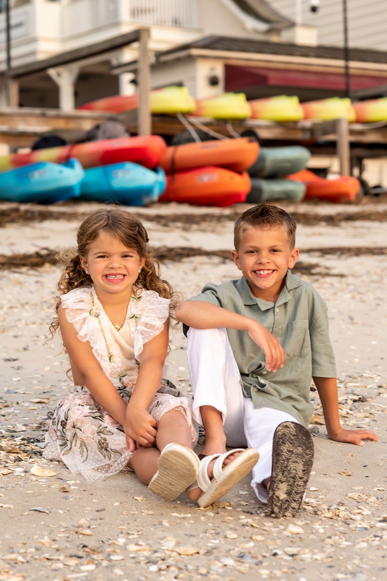 Two children lying casually on a sandy beach with colorful kayaks in the background.