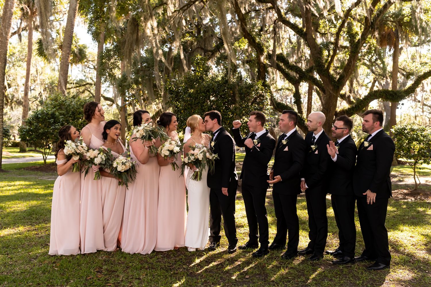 Wedding party poses in a line under sunlit trees wearing pink dresses and black suits during an outdoor ceremony.